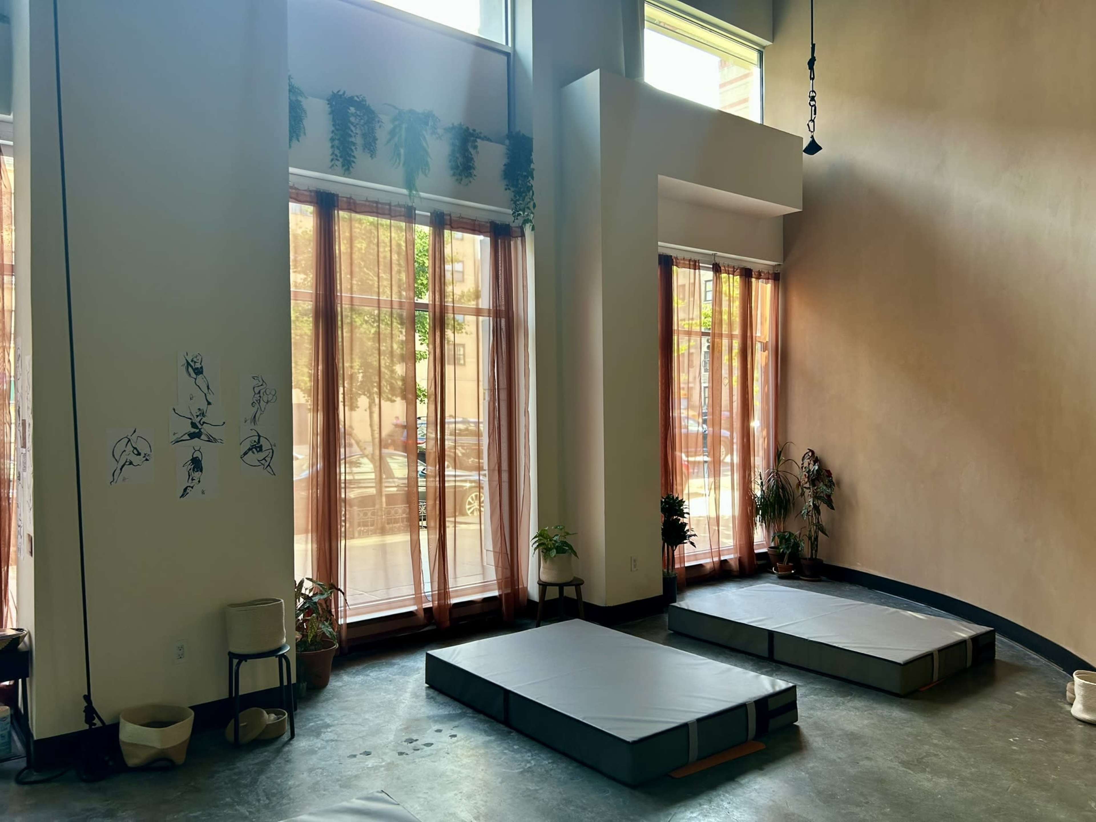 The image shows a spacious room with large windows, light curtains, and two simple mattresses positioned on a concrete floor.