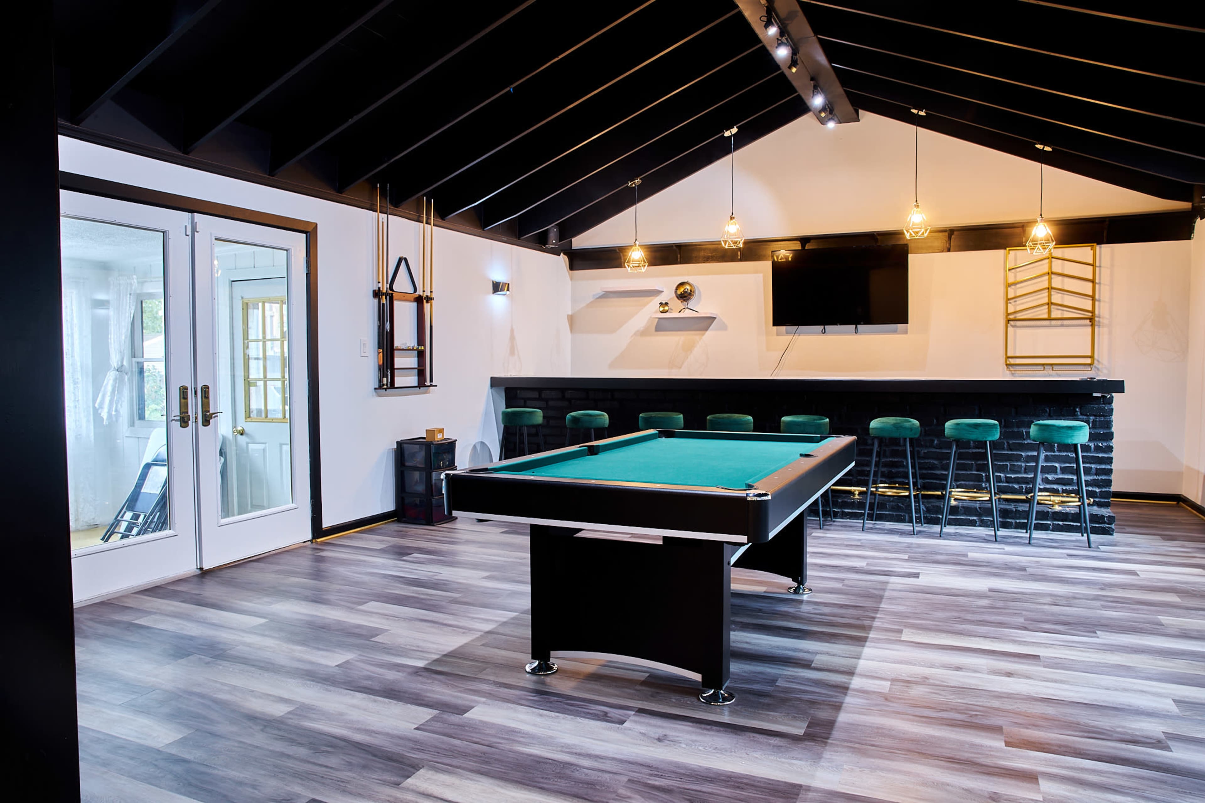 A modern game room featuring a billiards table, a bar counter with stools, and large windows allowing natural light.