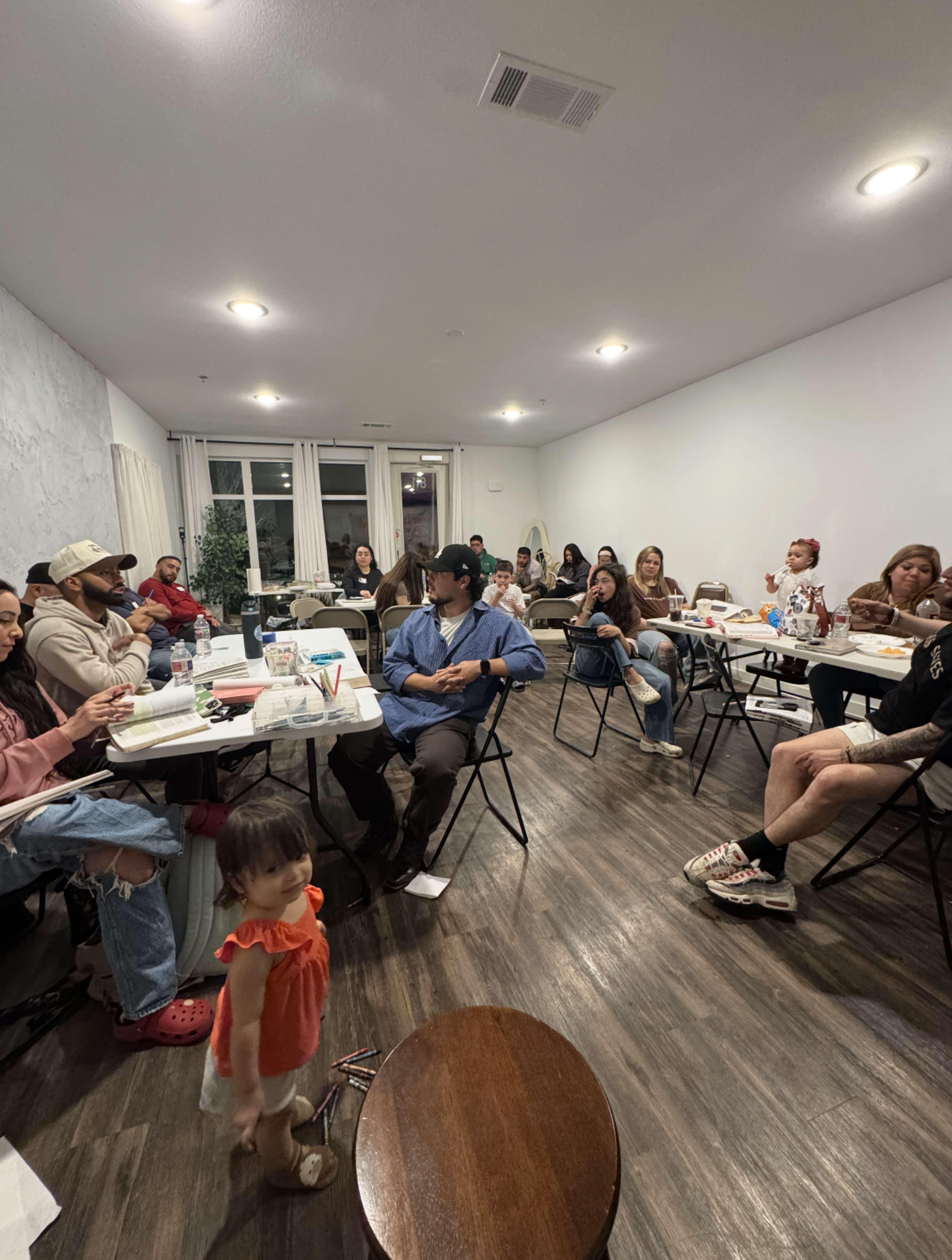A group of approximately twenty people sits in a well-lit room with tables and chairs, engaged in various activities, while a young child walks in the foreground.