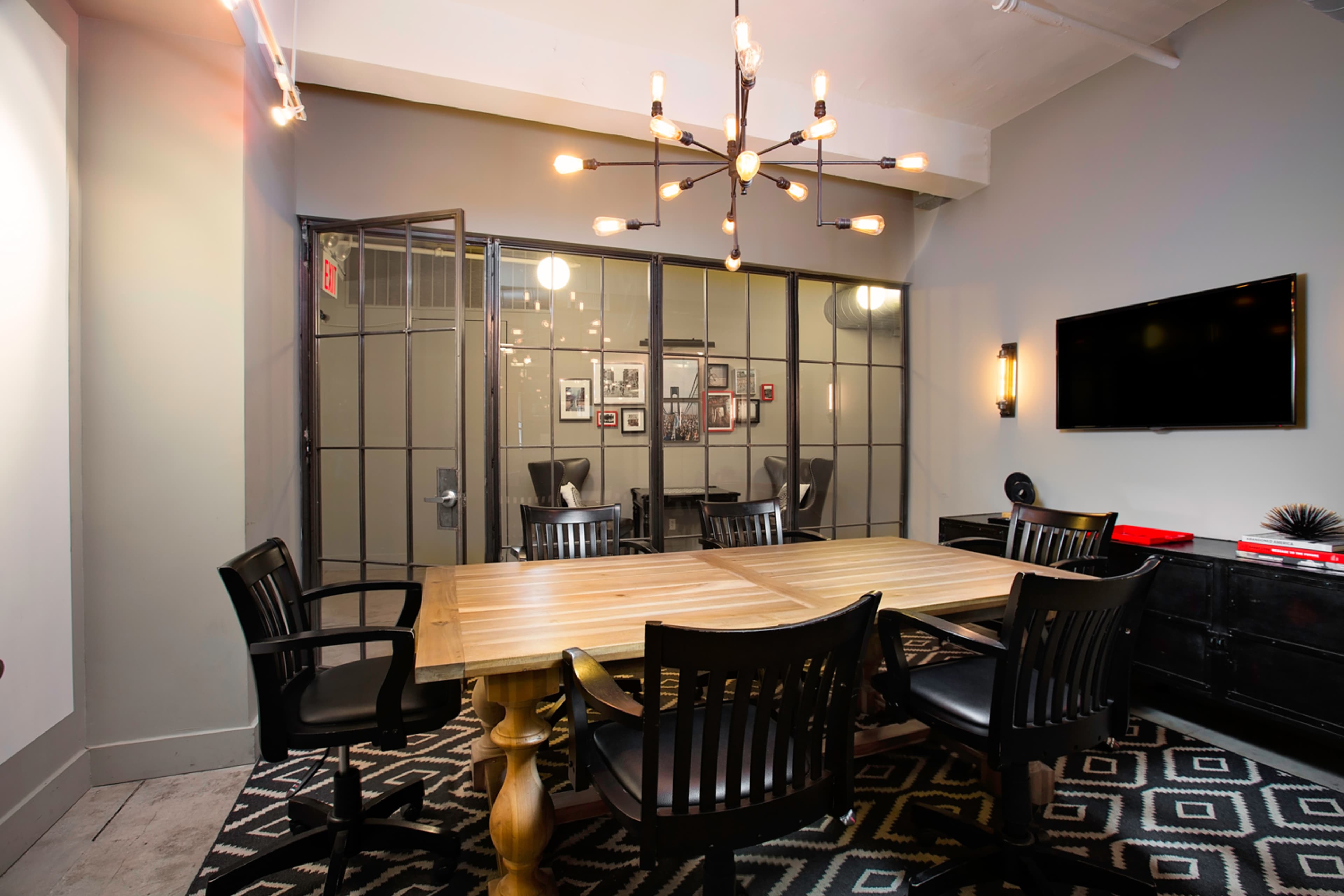 The image shows a modern conference room featuring a wooden table surrounded by black chairs, with a large glass door and wall of framed pictures in the background.