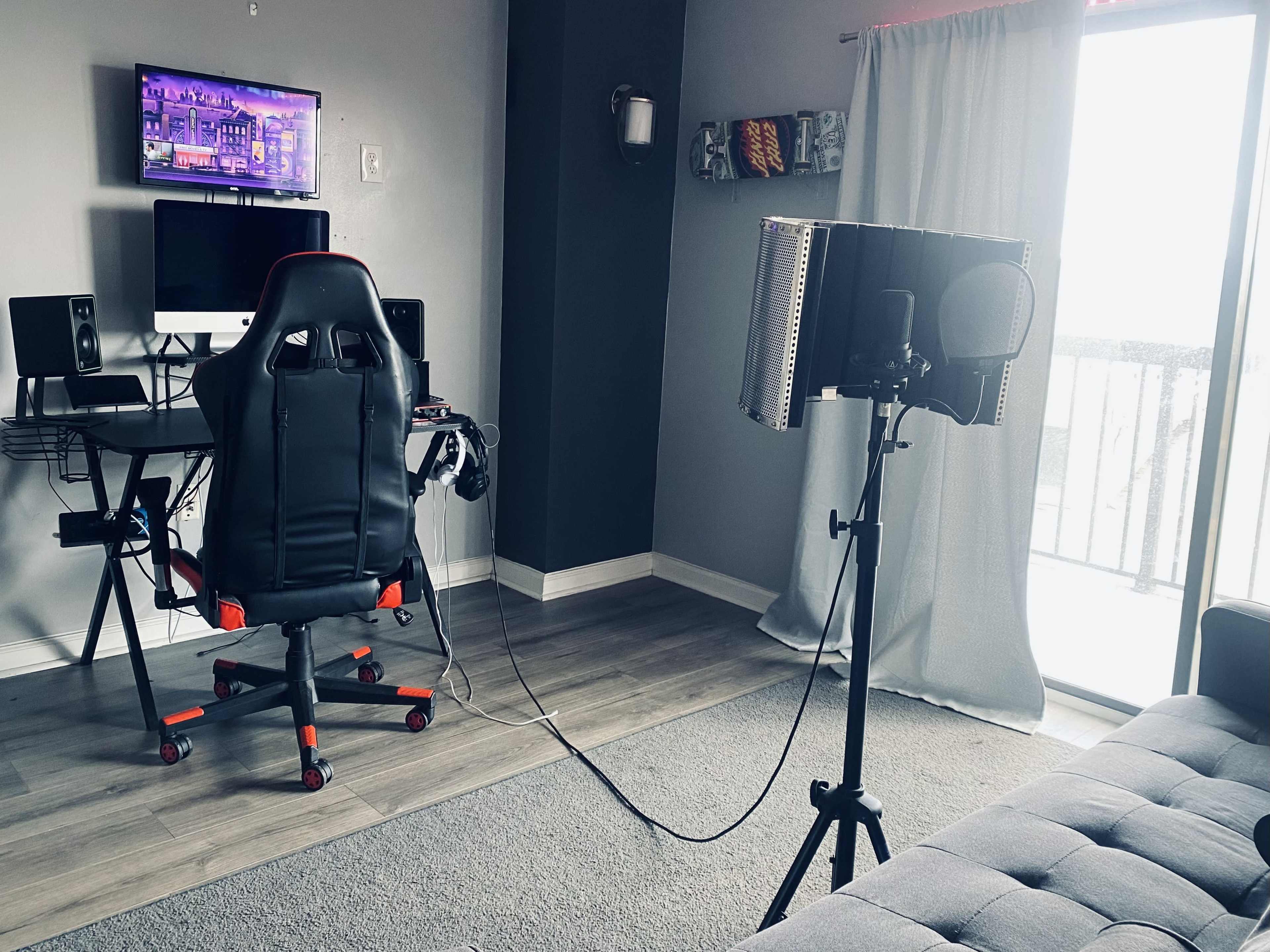 The image shows a modern workspace featuring a gaming chair, a desk with a computer monitor and speakers, and a microphone on a stand near a window.