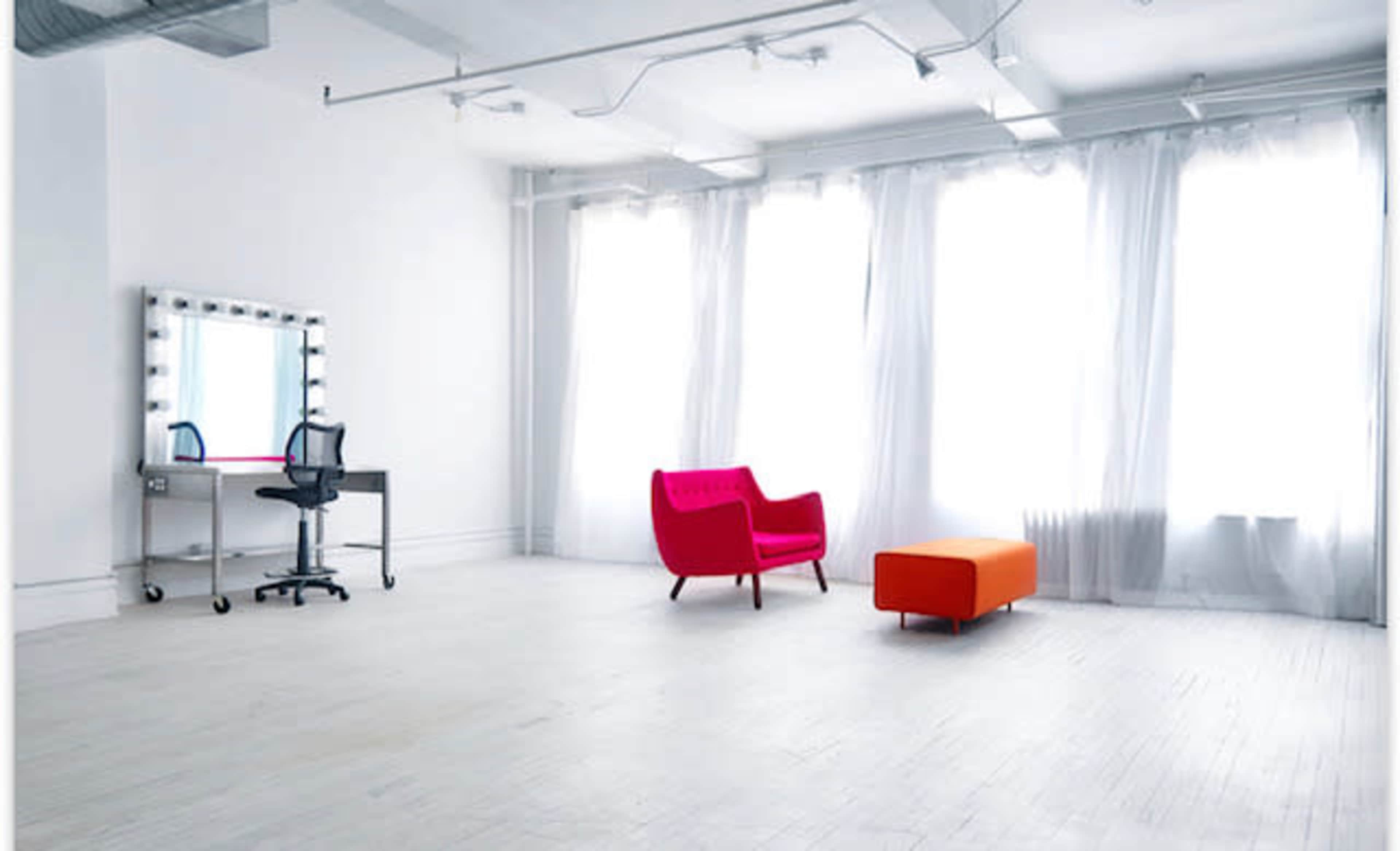 A spacious, well-lit room with large windows, a pink armchair, an orange bench, and a makeup vanity with lights.