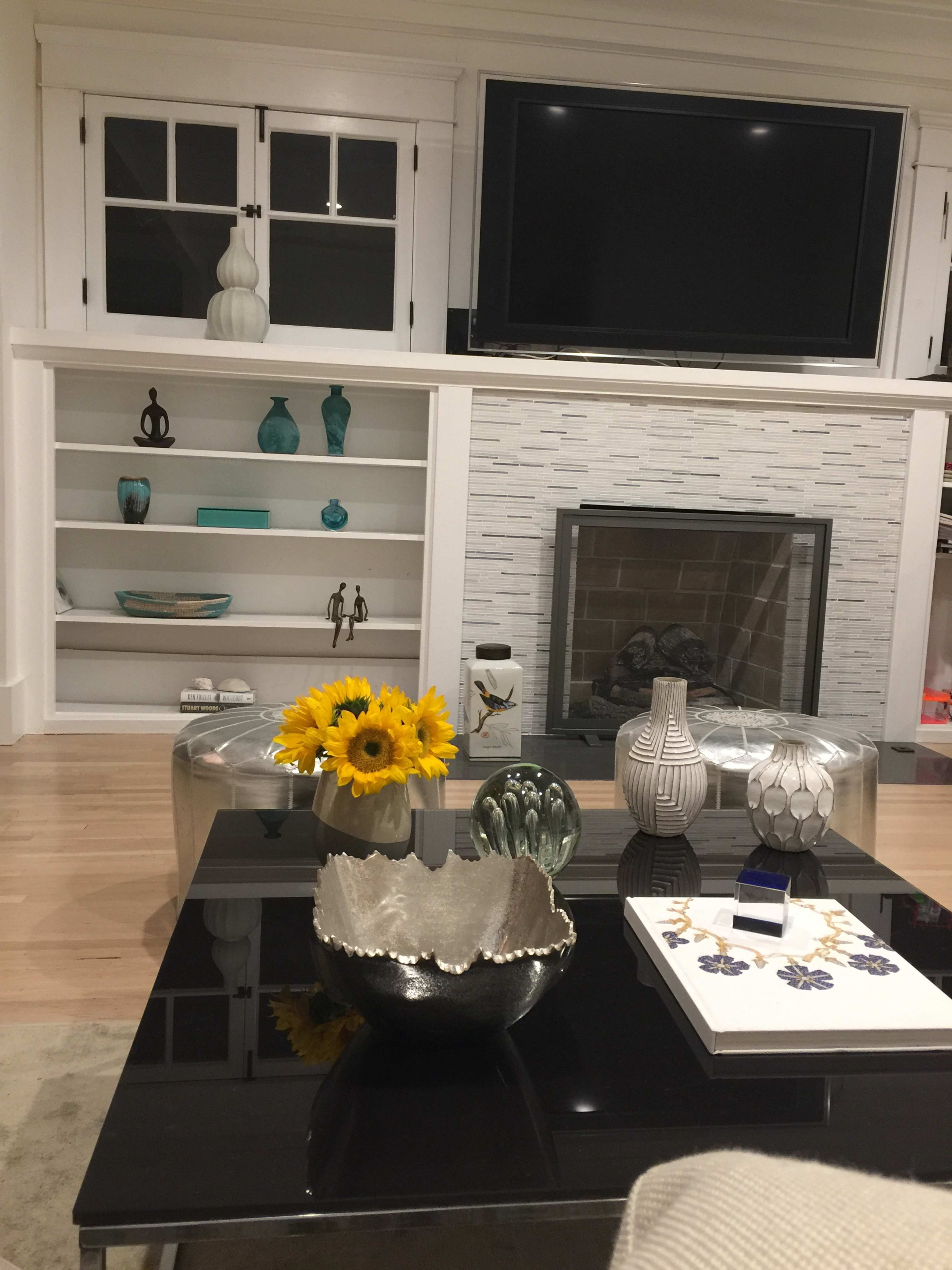 A living room with a television mounted on the wall, a fireplace with a stone surround, and a coffee table adorned with decorative items and a vase of sunflowers.