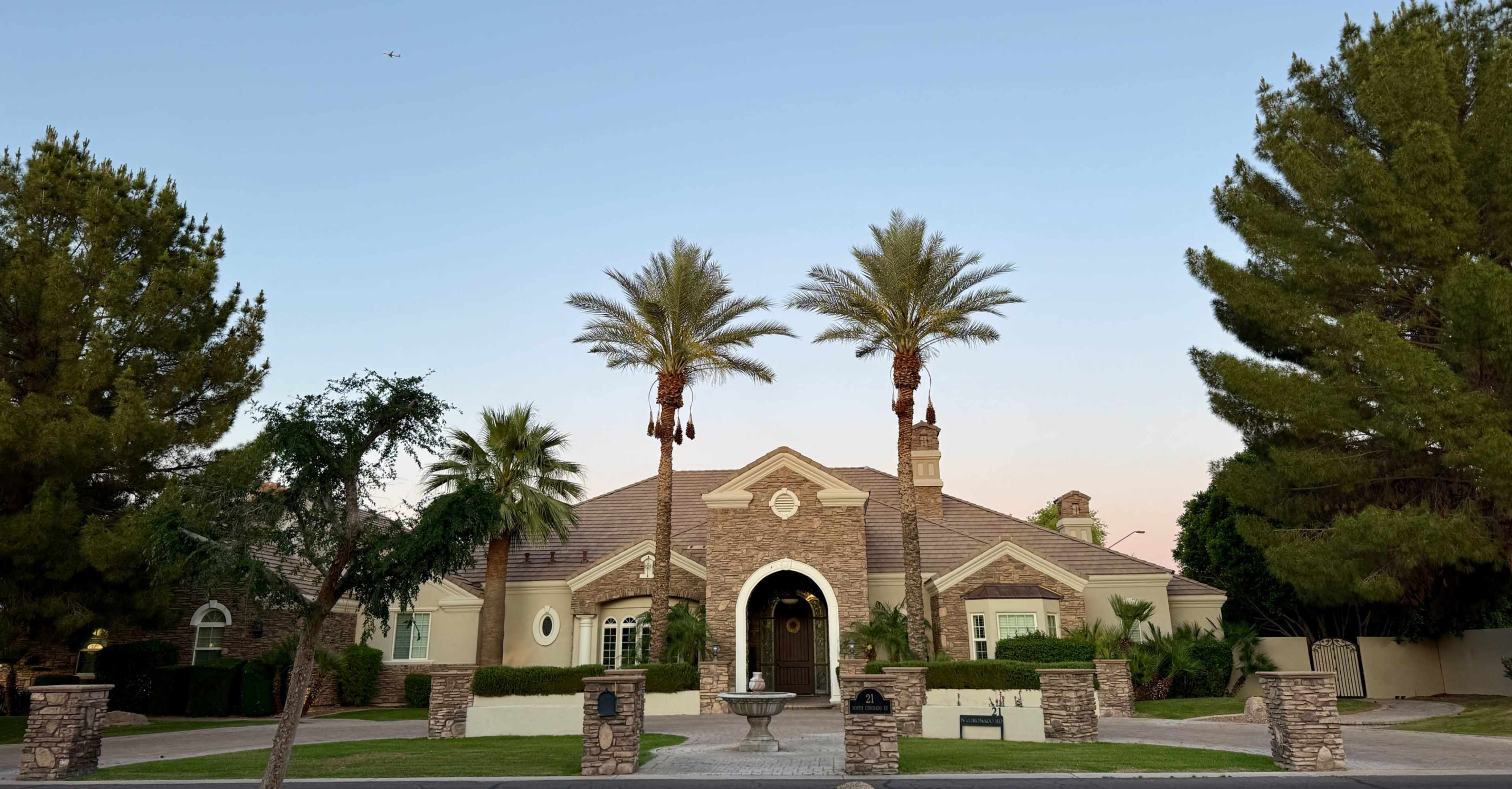 The image shows a large, single-story house surrounded by palm trees and well-manicured landscaping.