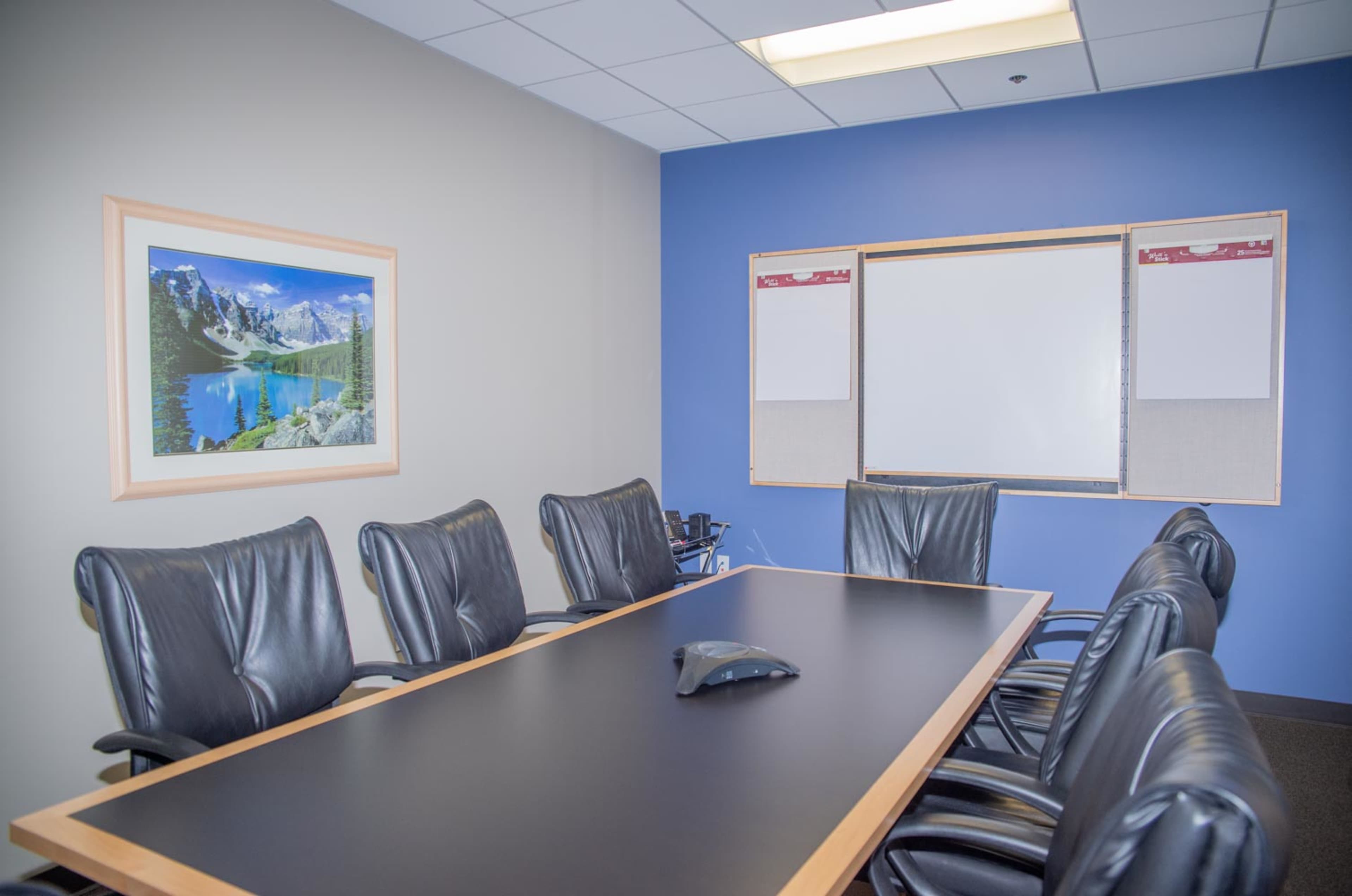 A conference room with a long black table surrounded by eight black leather chairs, a whiteboard on one wall, and a landscape photograph on another wall.