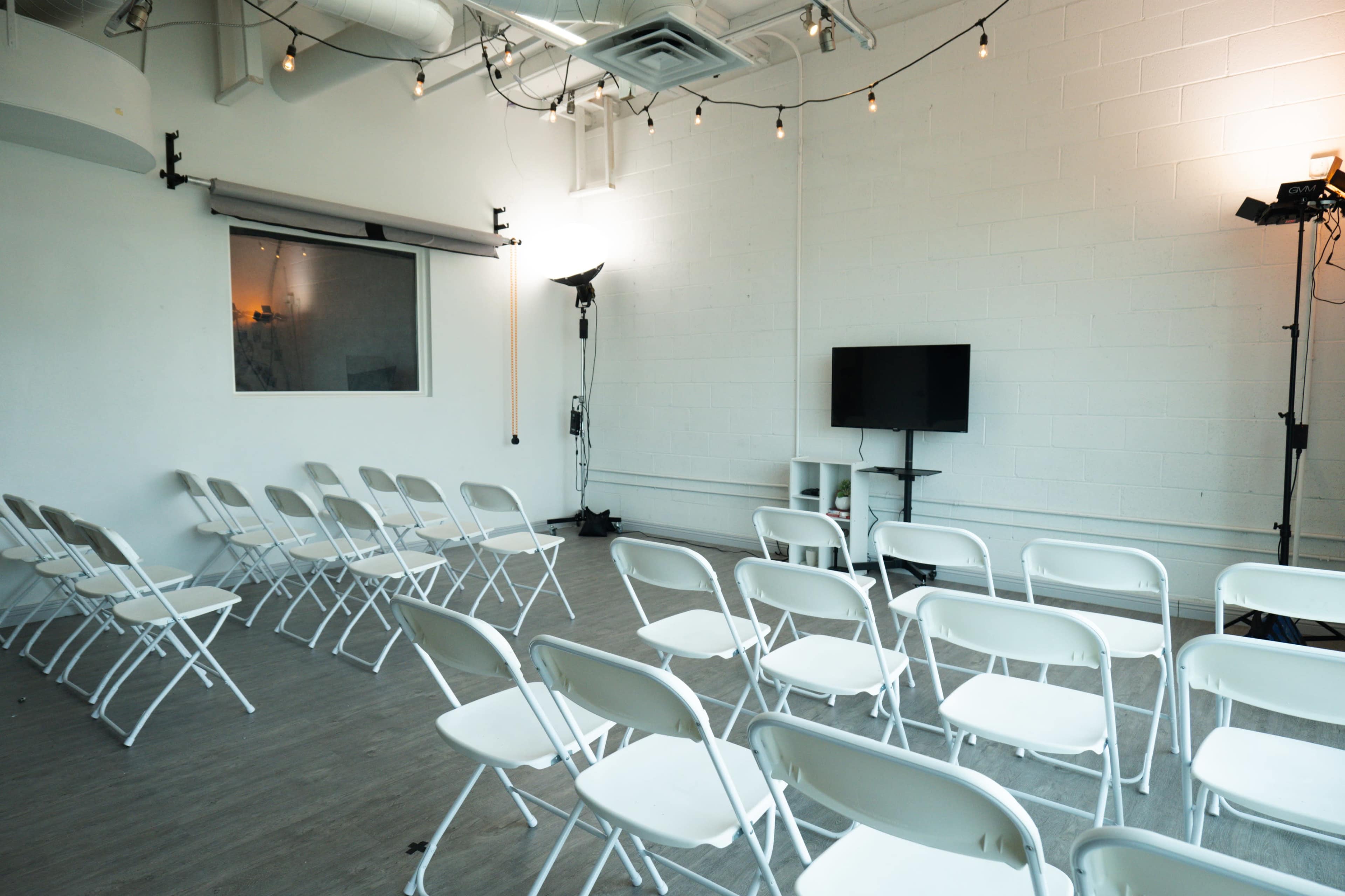 A row of white folding chairs are arranged in a brightly lit room with a television and lighting equipment set up at the front.