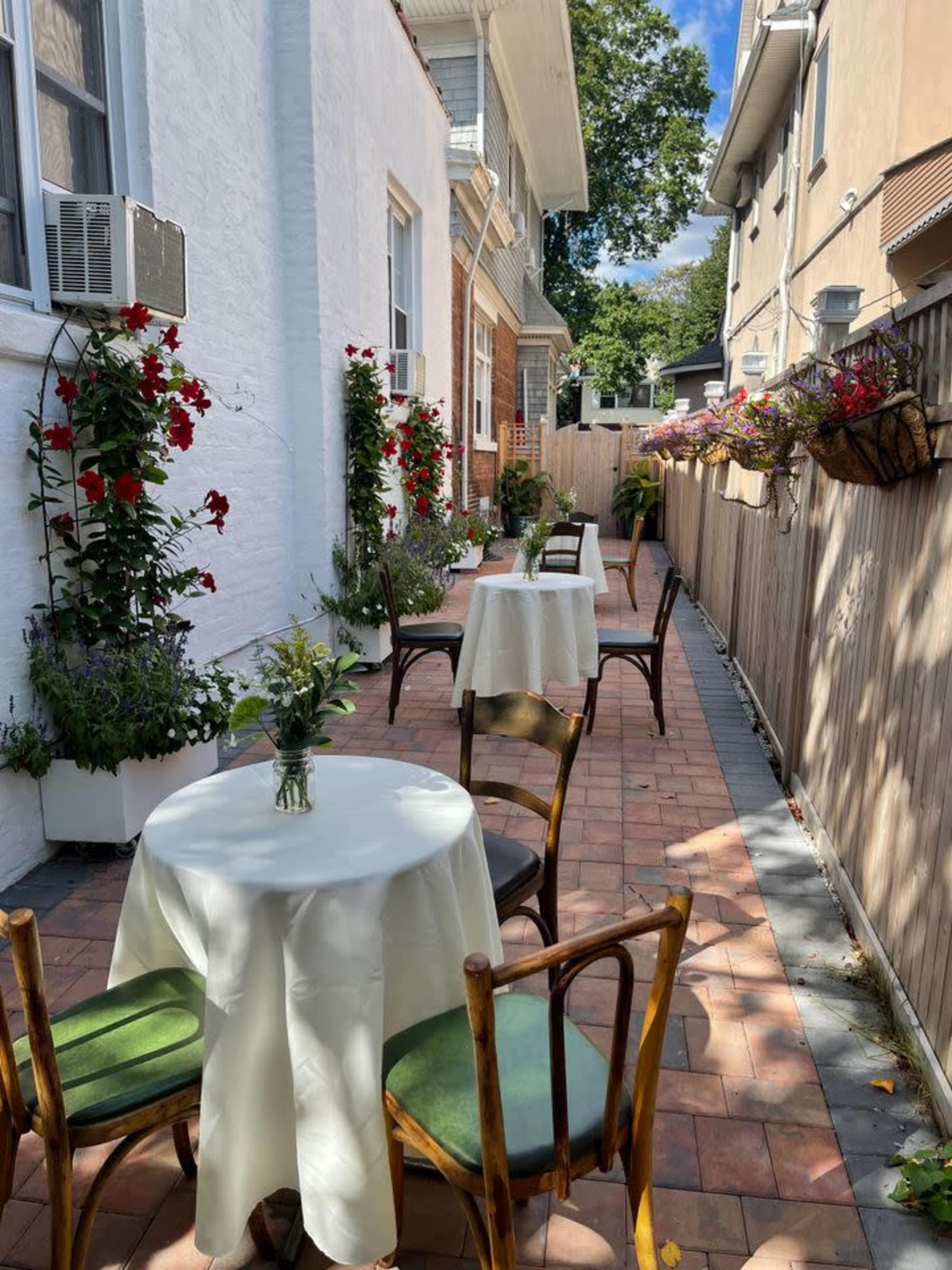 A narrow outdoor patio is lined with tables covered in white tablecloths, surrounded by potted flowers and a wooden fence.