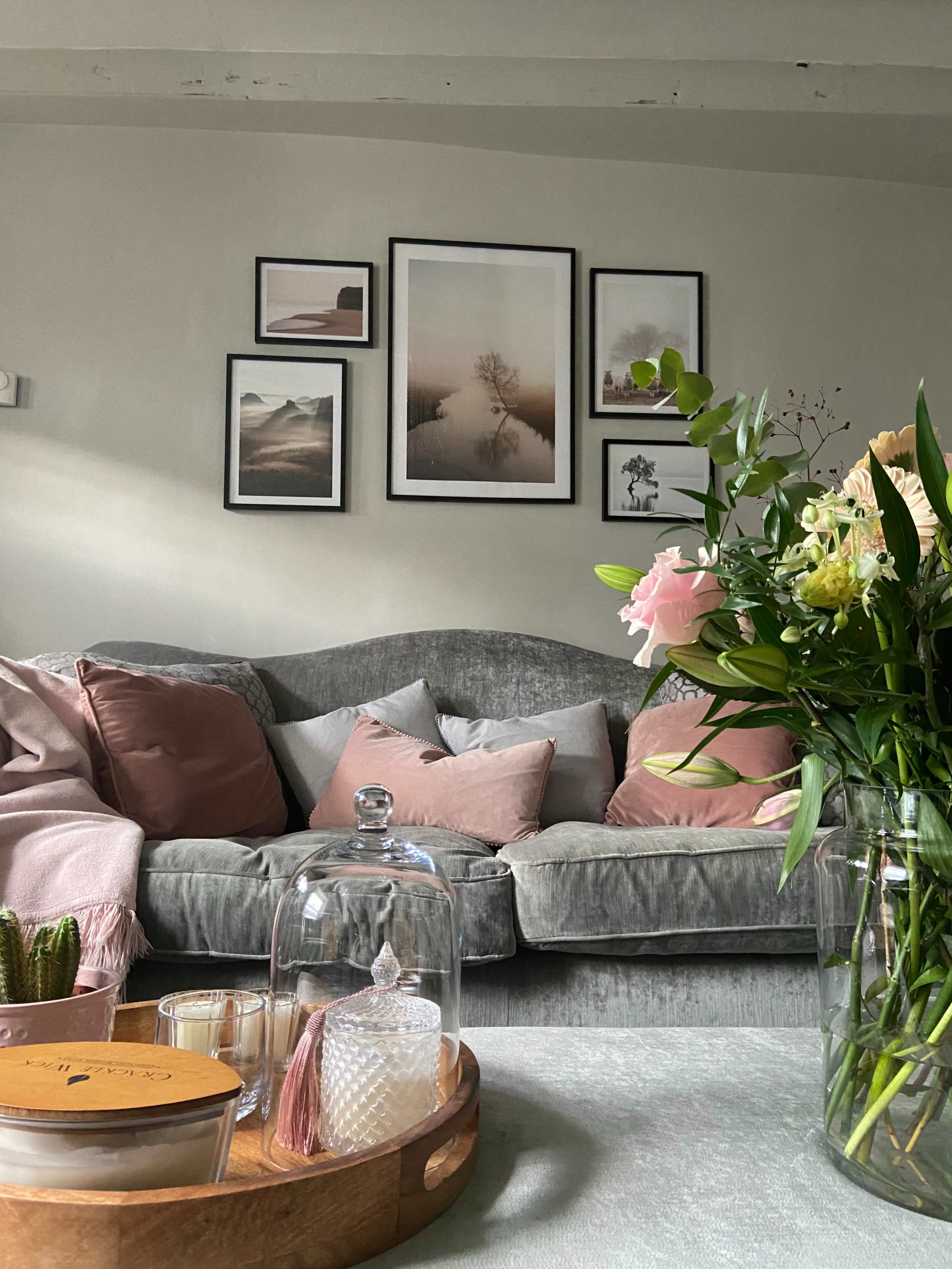 A cozy living room features a gray sofa adorned with pink cushions, framed photographs on the wall, and a coffee table with decorative items and a vase of flowers.