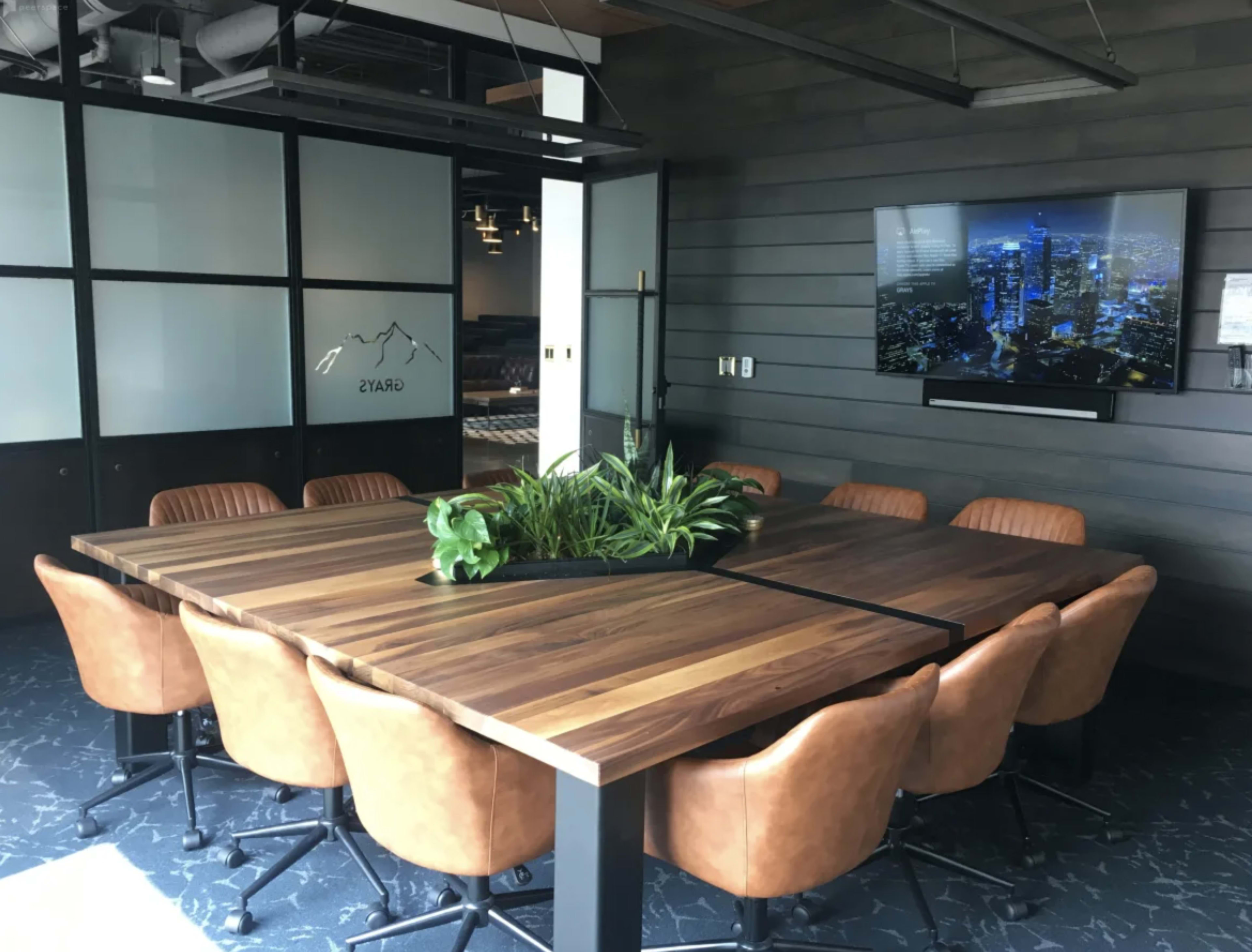 A large wooden conference table surrounded by brown leather chairs is positioned in a modern meeting room with a television mounted on the wall.