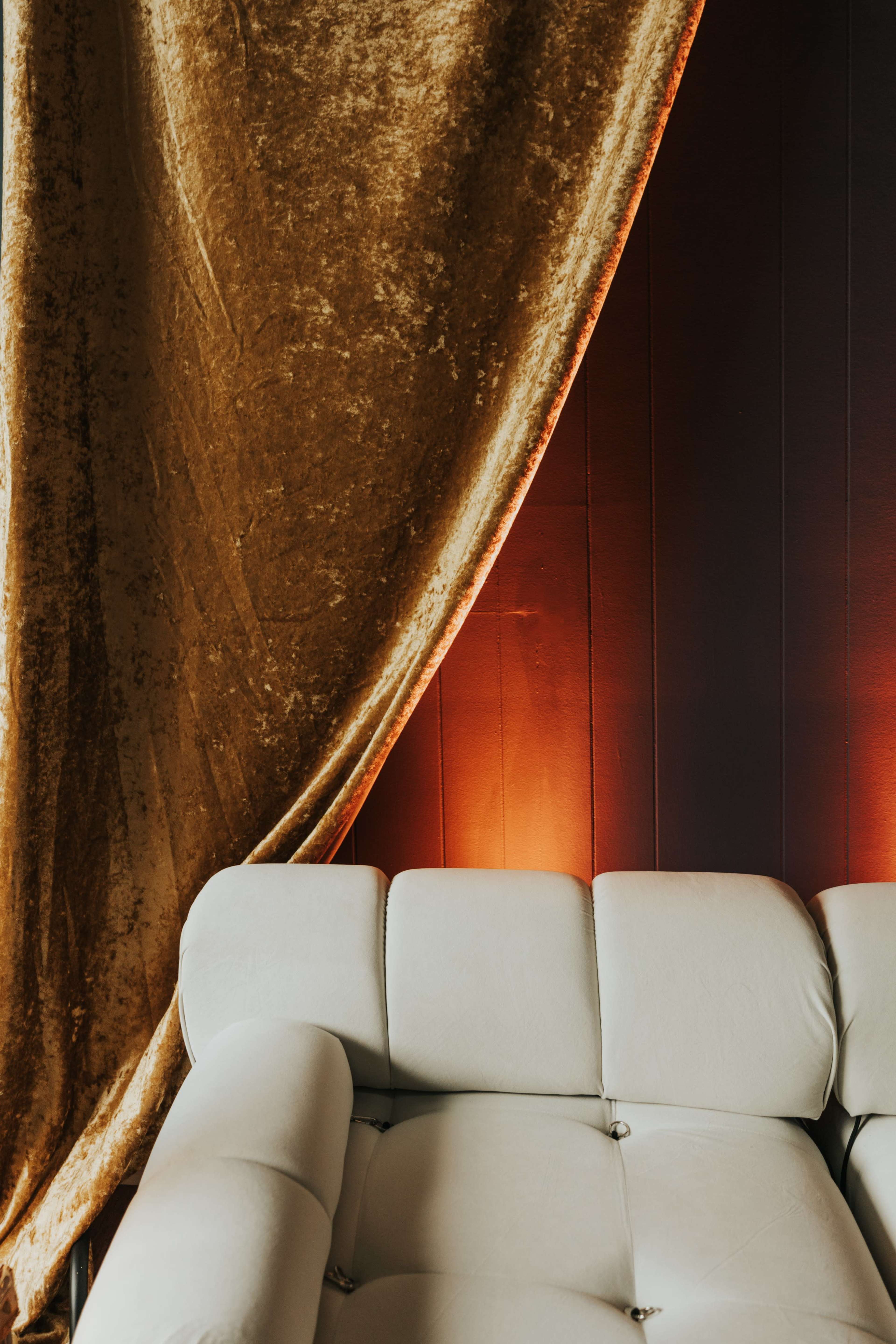 A light-colored sofa partly obscured by a gold velvet curtain against a dark wall with ambient lighting.