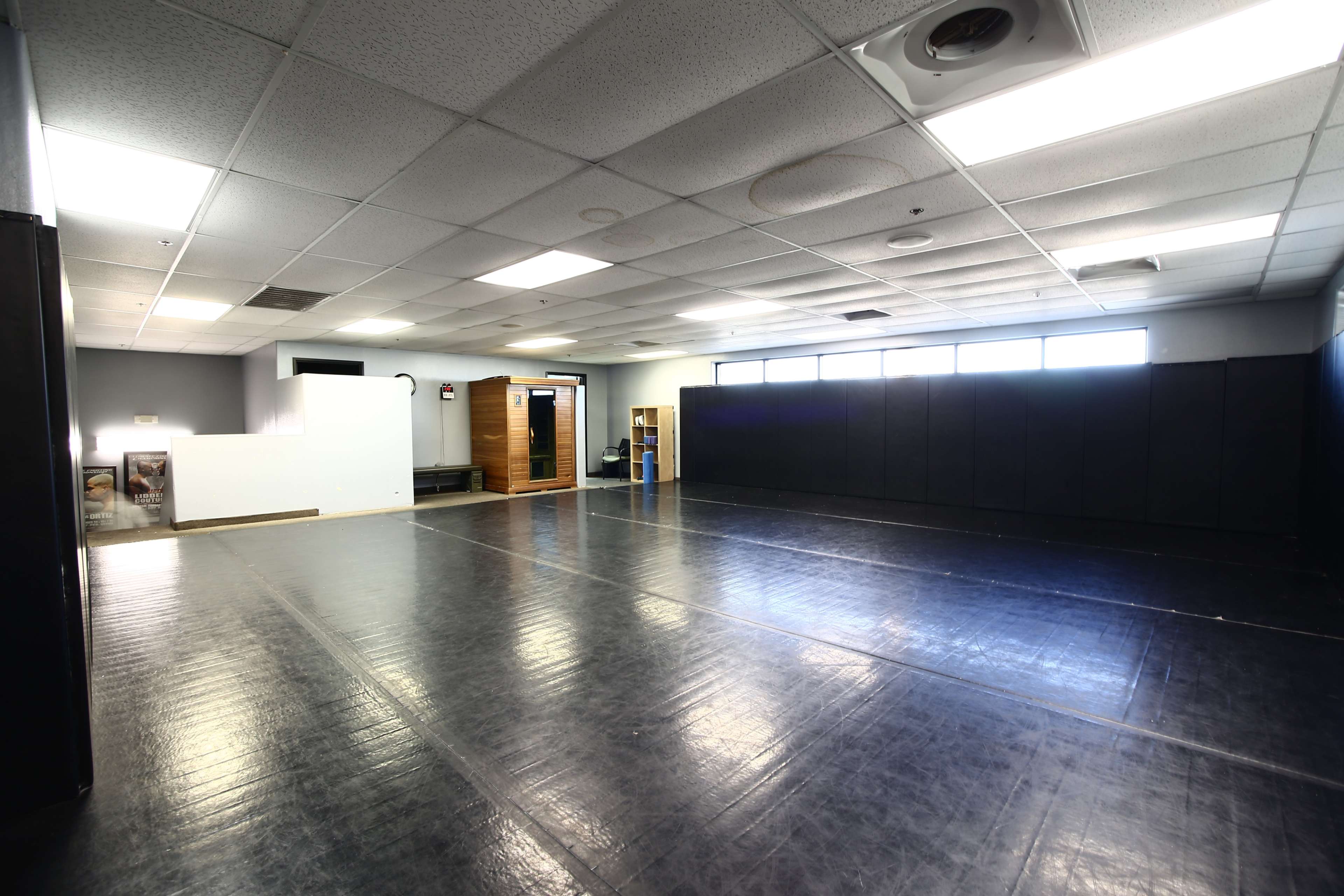 A large, empty room with black mats on the floor and padded walls, featuring bright overhead lights and large windows.
