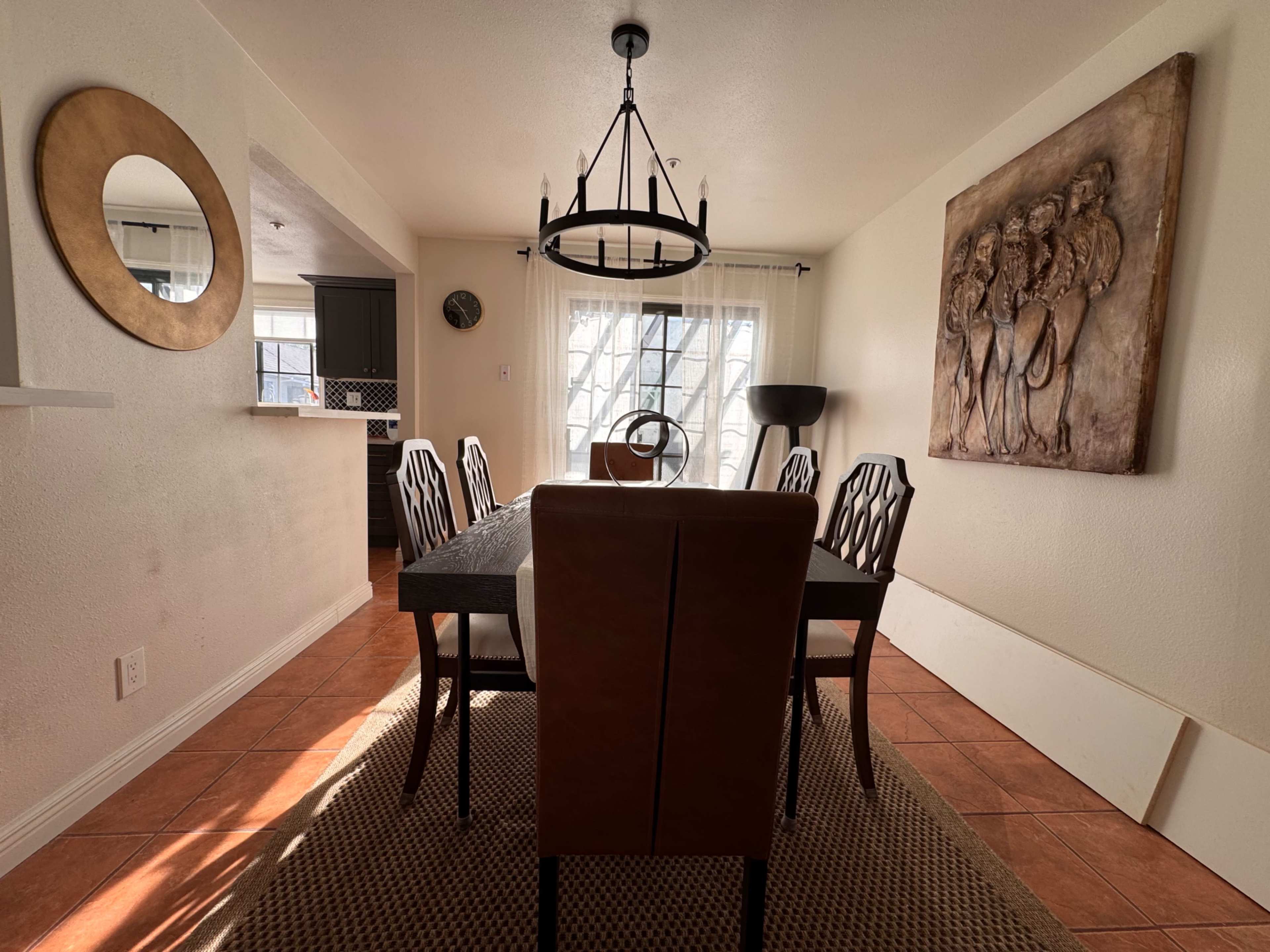 The image shows a dining room with a rectangular table surrounded by chairs, a chandelier overhead, and wall decor featuring a relief sculpture.