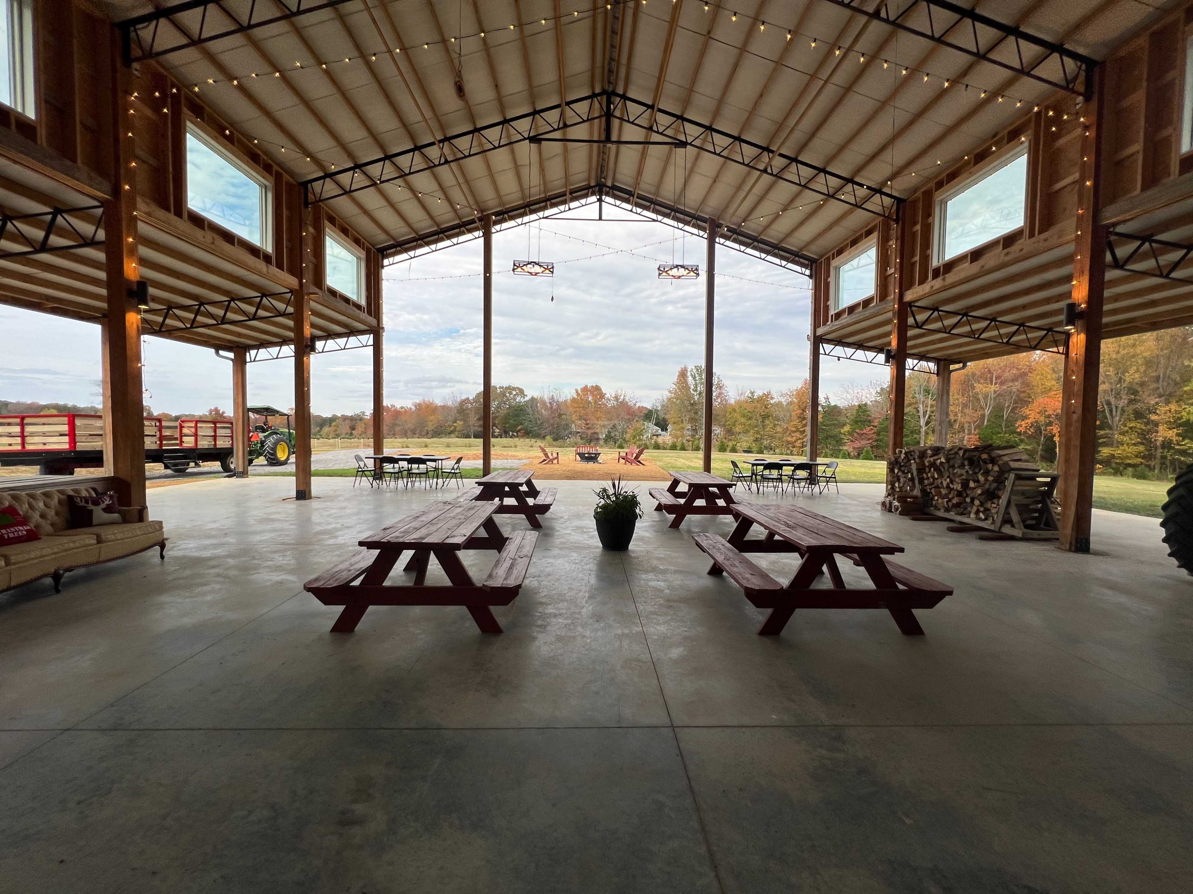 A spacious barn interior with wooden picnic tables, large windows, and a view of a landscaped outdoor area with a tractor and firewood stacks.