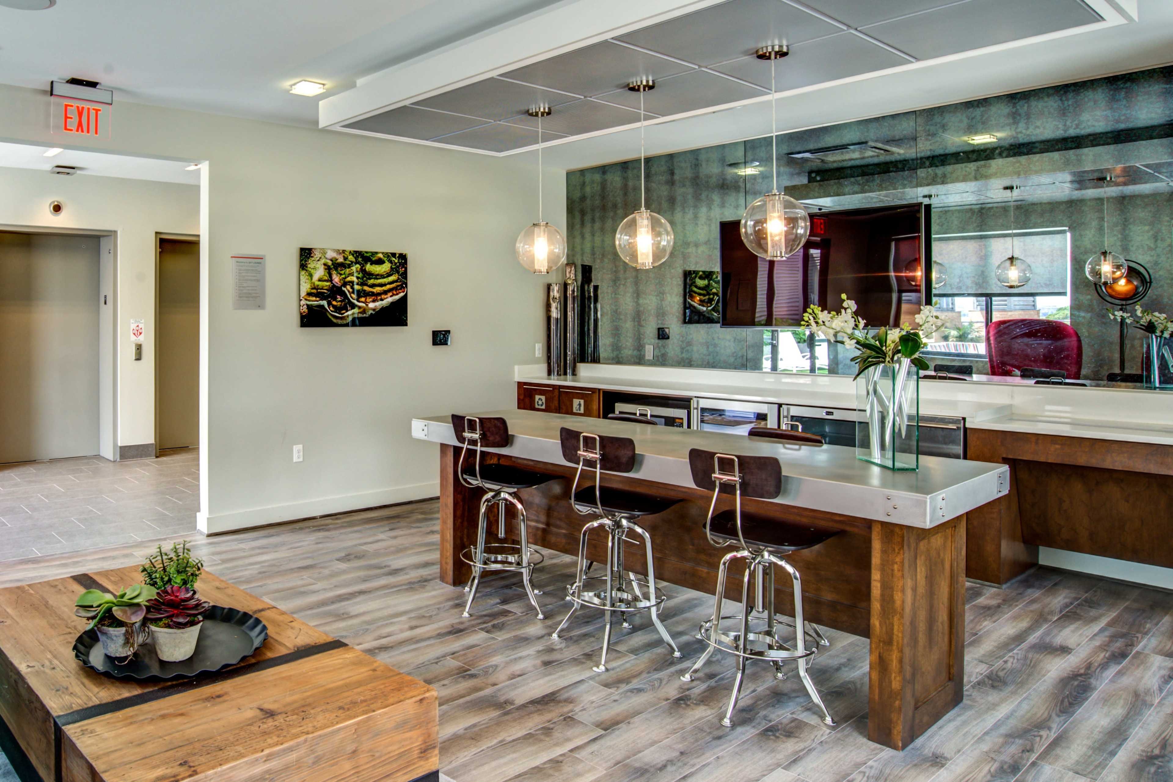 The image shows a modern kitchen and dining area featuring a long countertop with bar stools, a large mirror, and a wooden table with decorative plants.