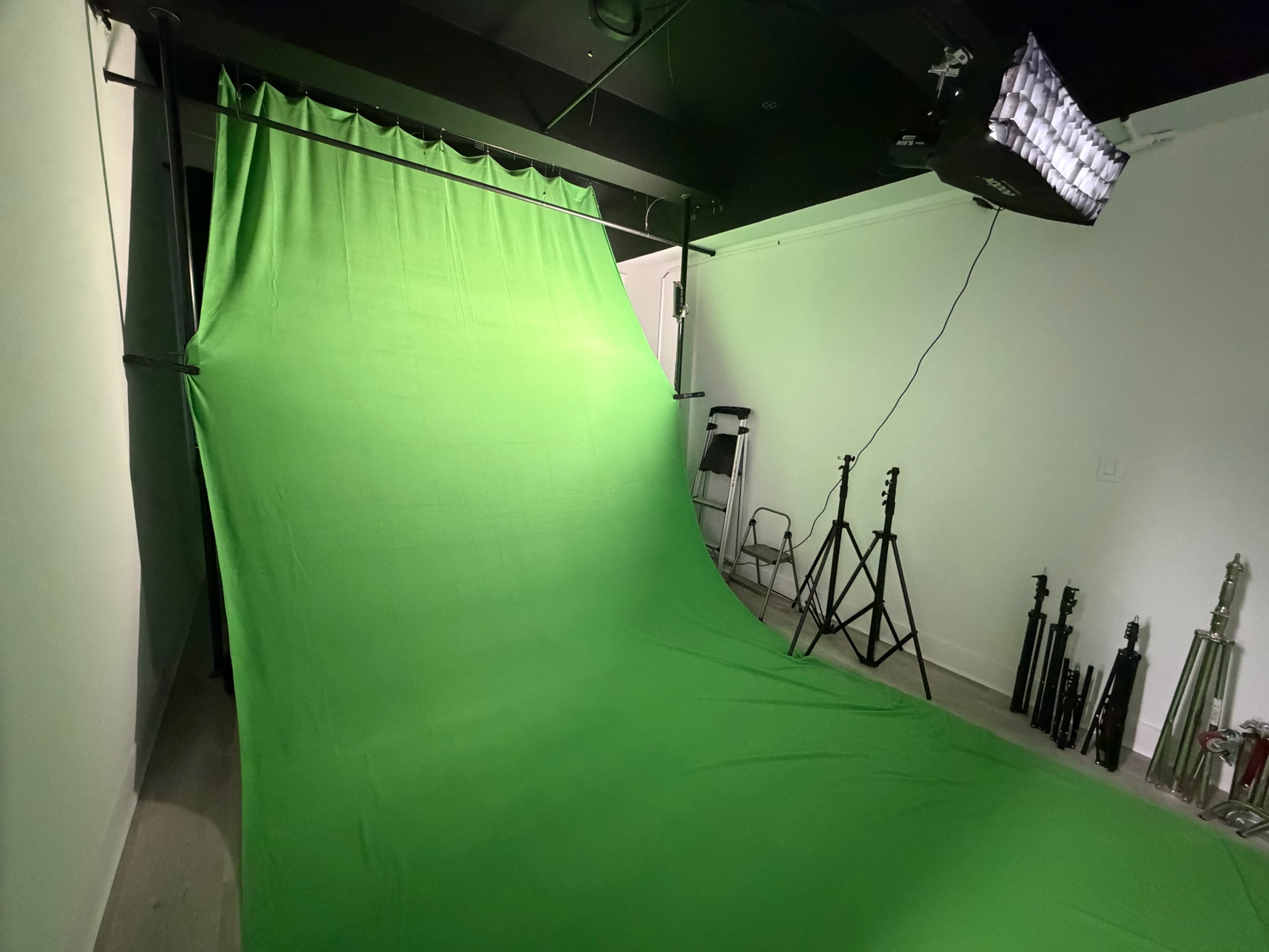 The image shows a photography studio with a large green backdrop, studio lights, and various equipment including stands and a chair.