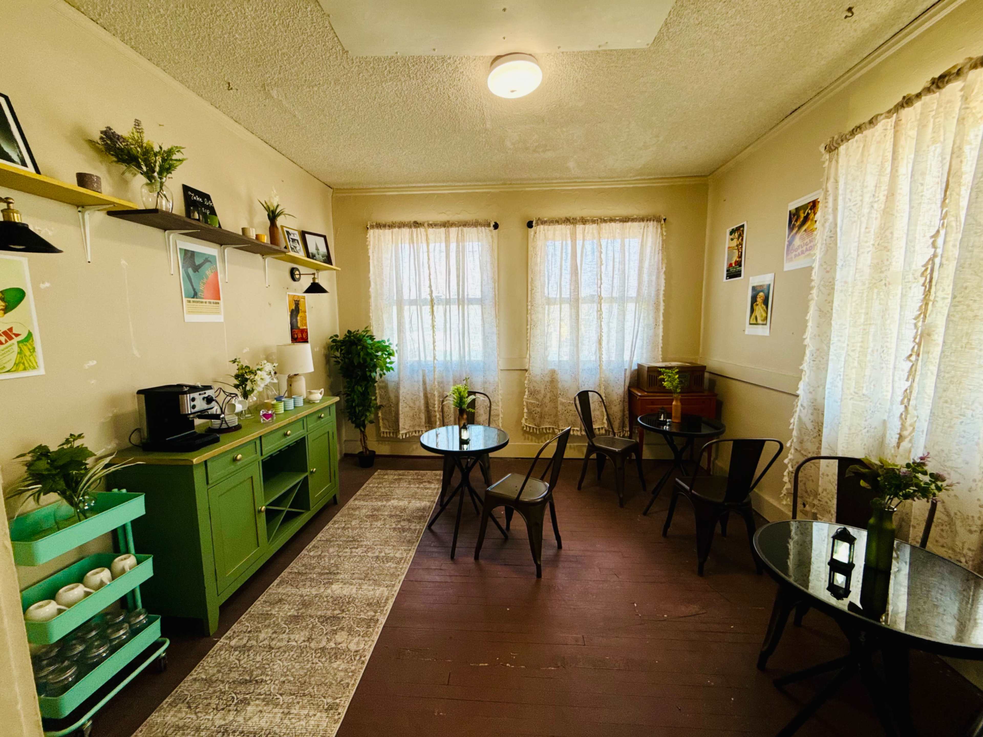 The room features a cozy seating area with four small tables, a green cabinet, and large windows covered by lace curtains.