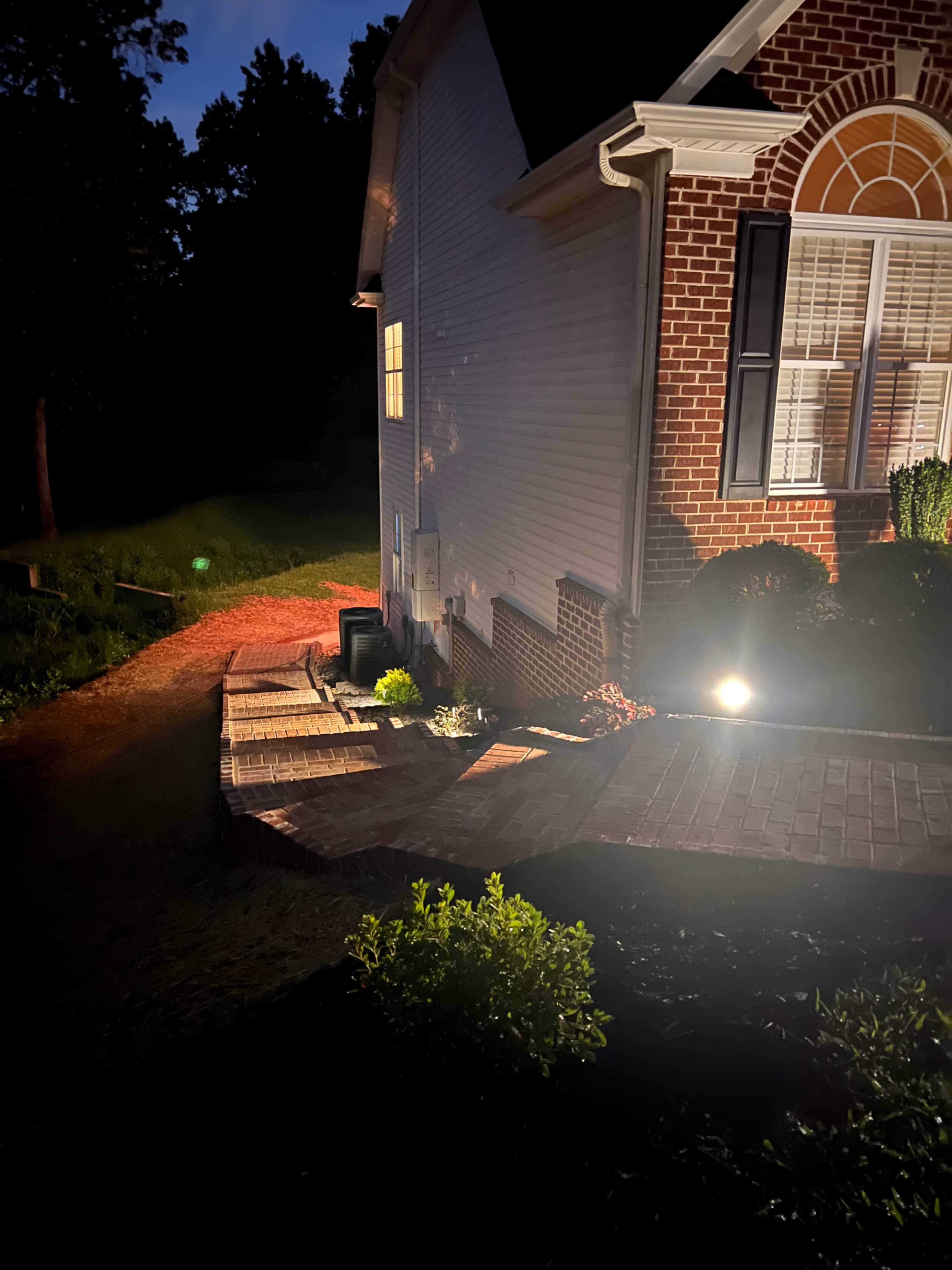 A well-lit pathway leading to a brick house at night, with landscaped greenery along the edges.