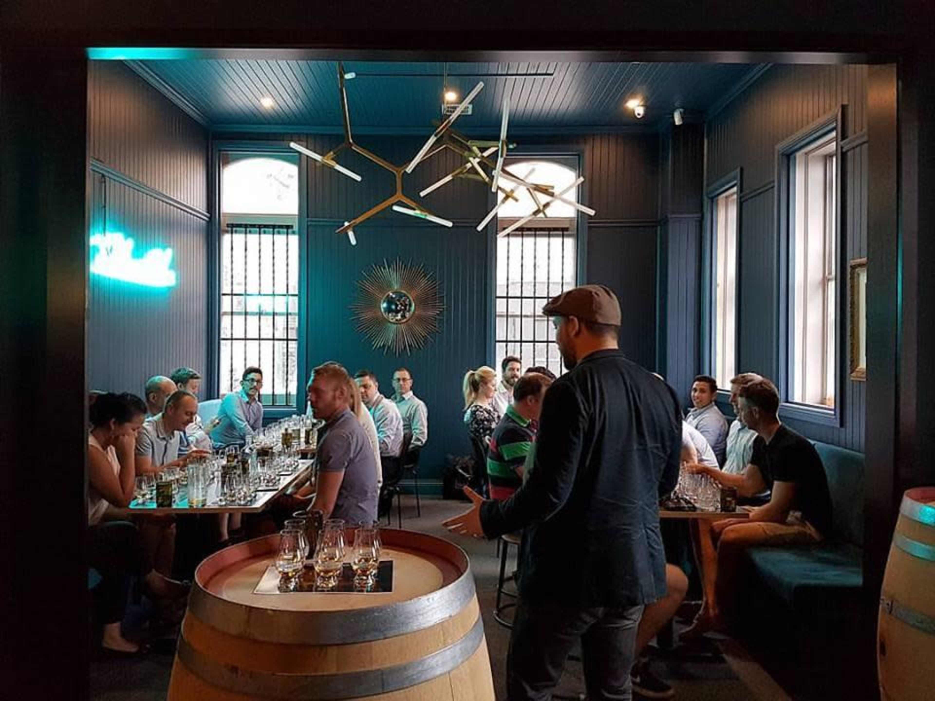 A group of people sits around tables in a dimly lit room with large windows and stylish decor, while one man stands near a barrel in the foreground.