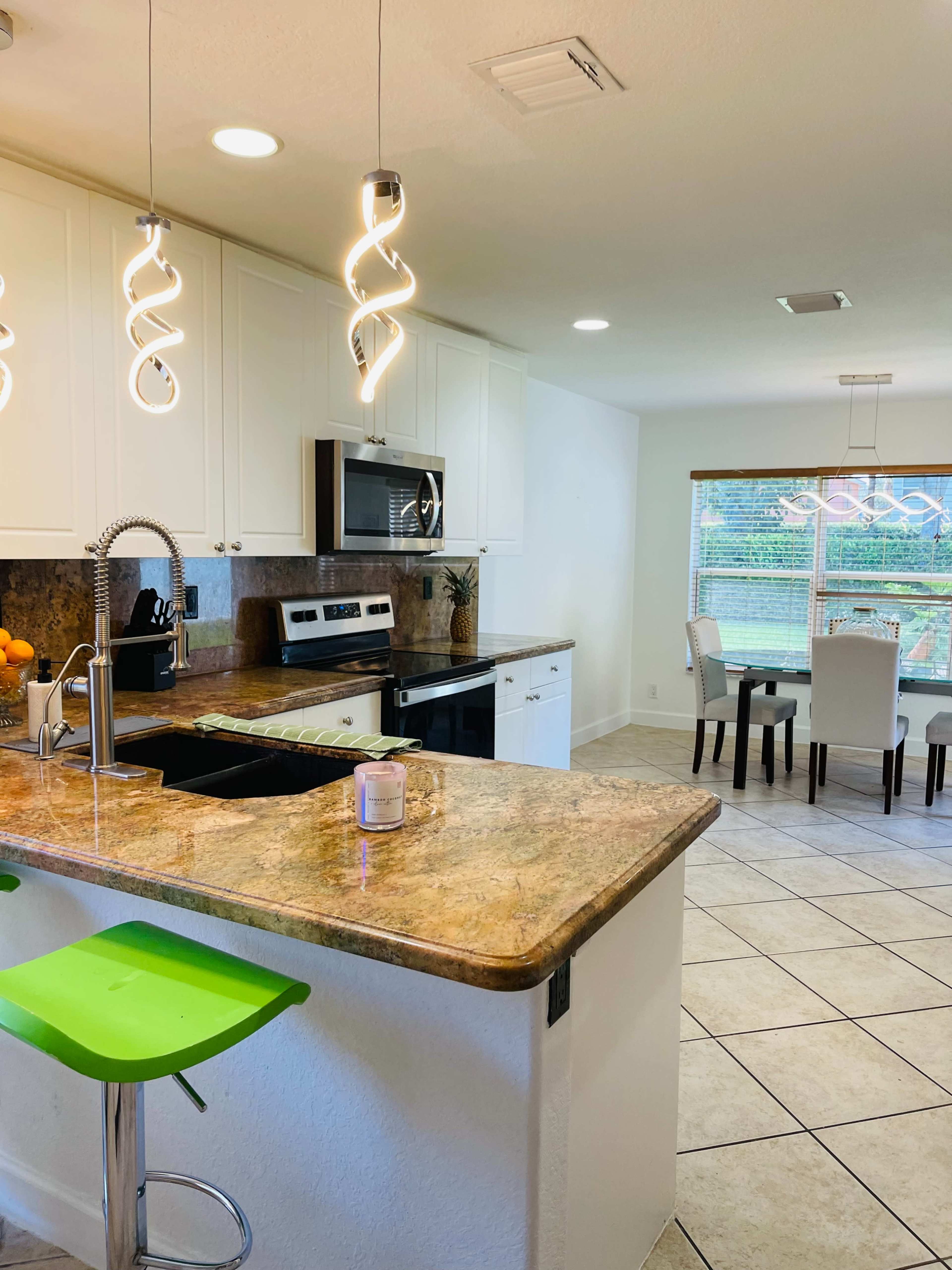 A modern kitchen features a granite countertop with bar stools, stainless steel appliances, and a dining area with a table and chairs, illuminated by pendant lights.