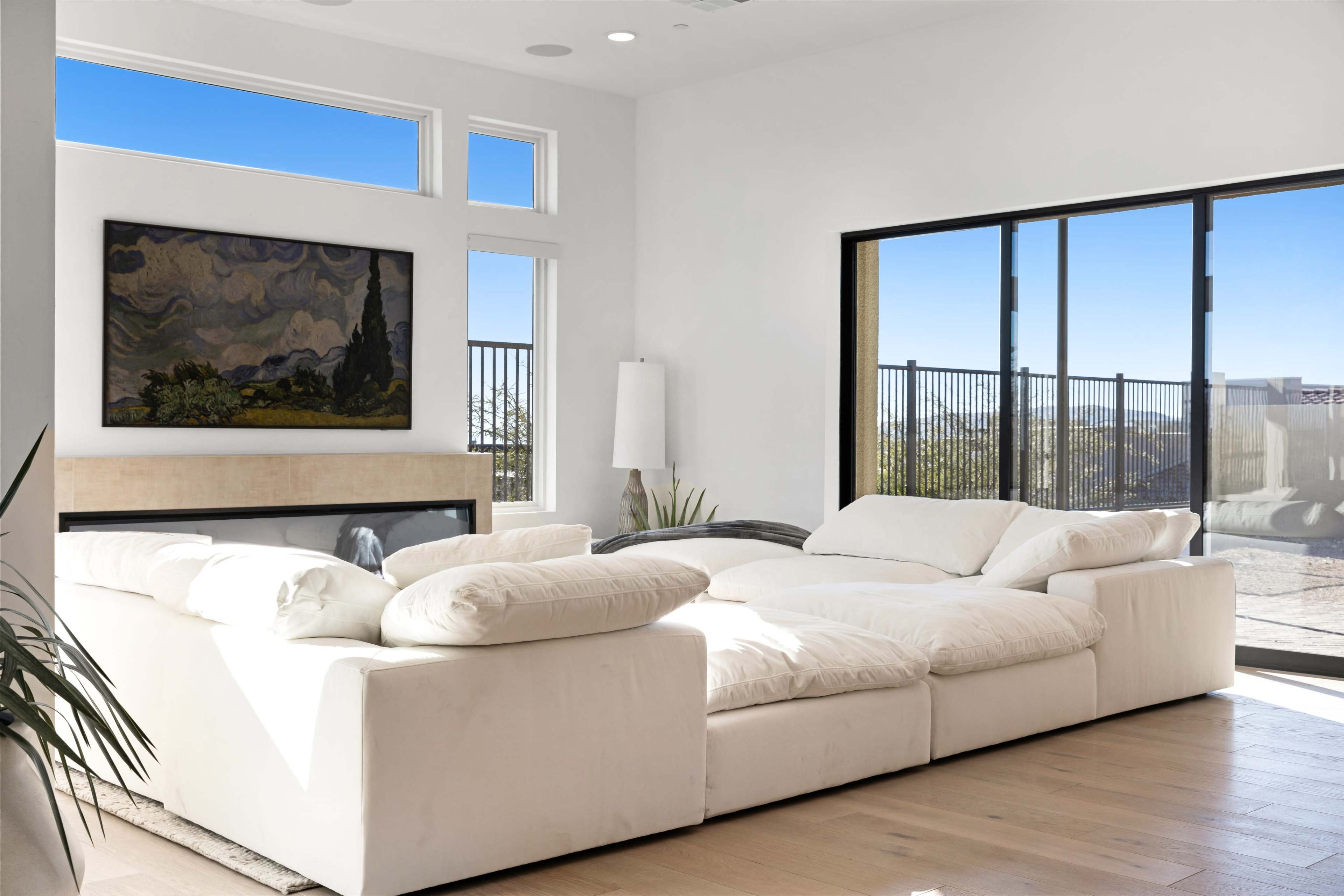 A modern living room features a large white sectional sofa, a flat-screen TV, and a painting on the wall, with large windows providing a view of the outdoors.
