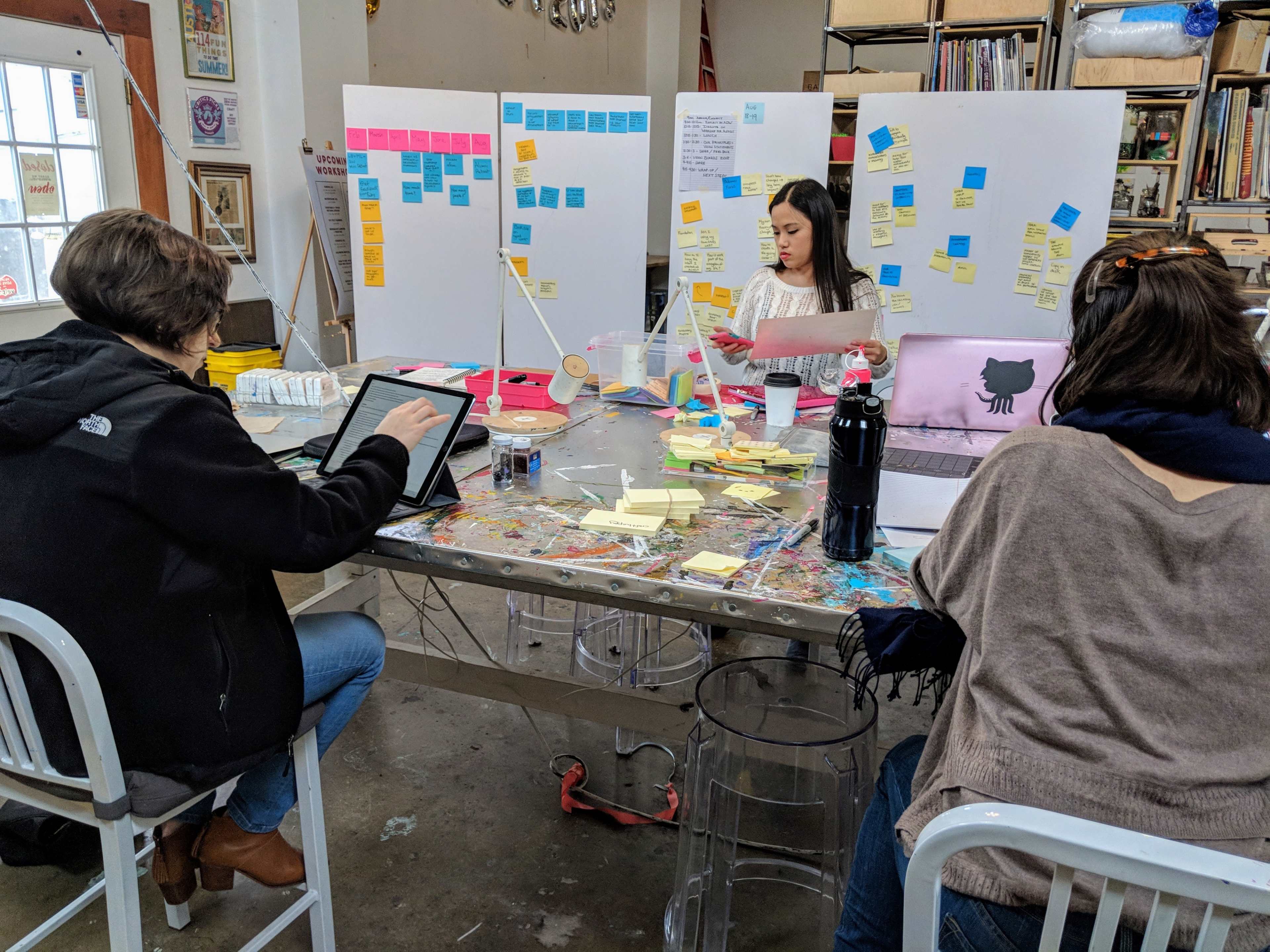 Three people collaborate in a studio filled with sticky notes on boards and a cluttered table covered in various materials.