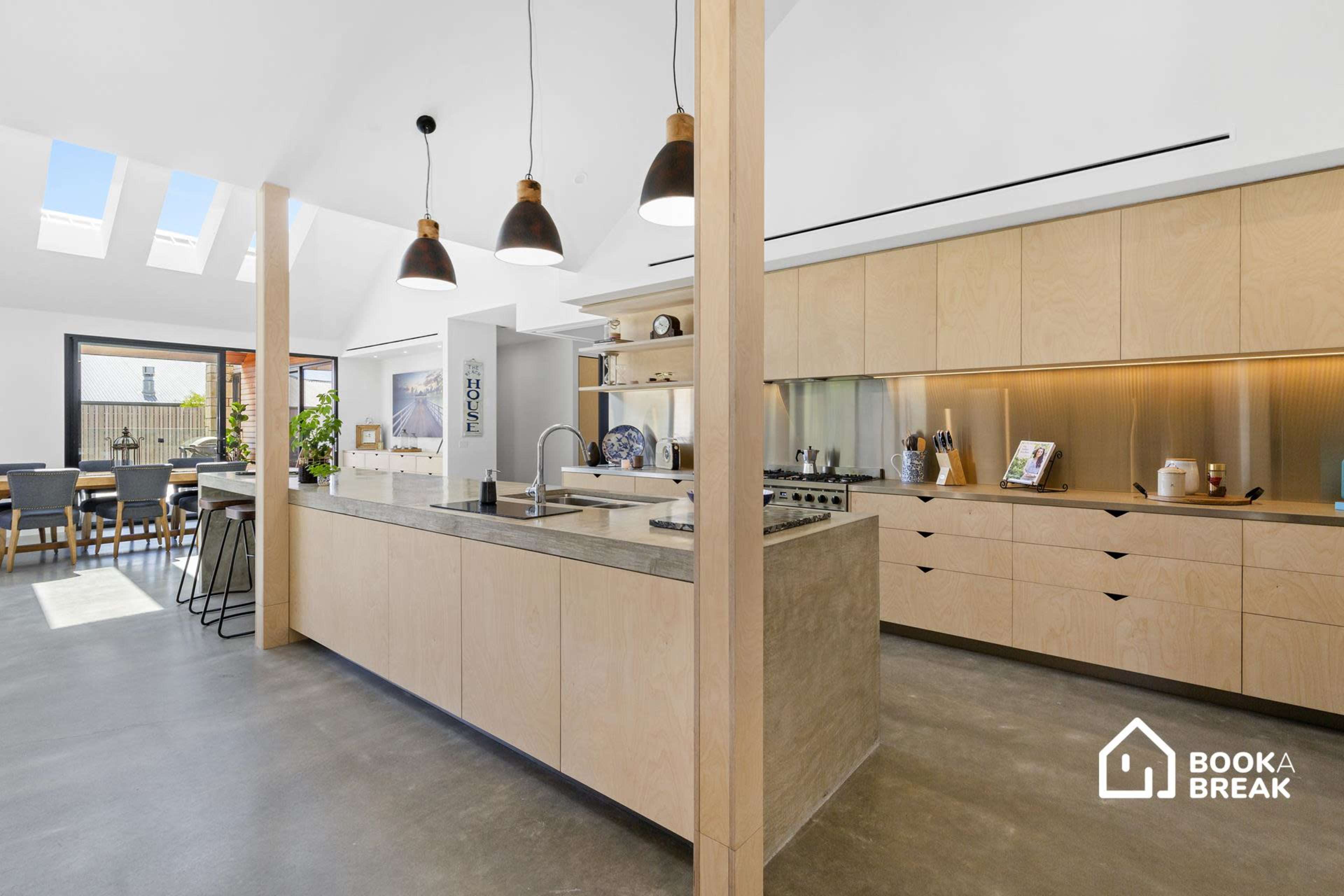 A modern kitchen features a central island with a concrete countertop, wooden cabinetry, and large windows that allow natural light to fill the space.