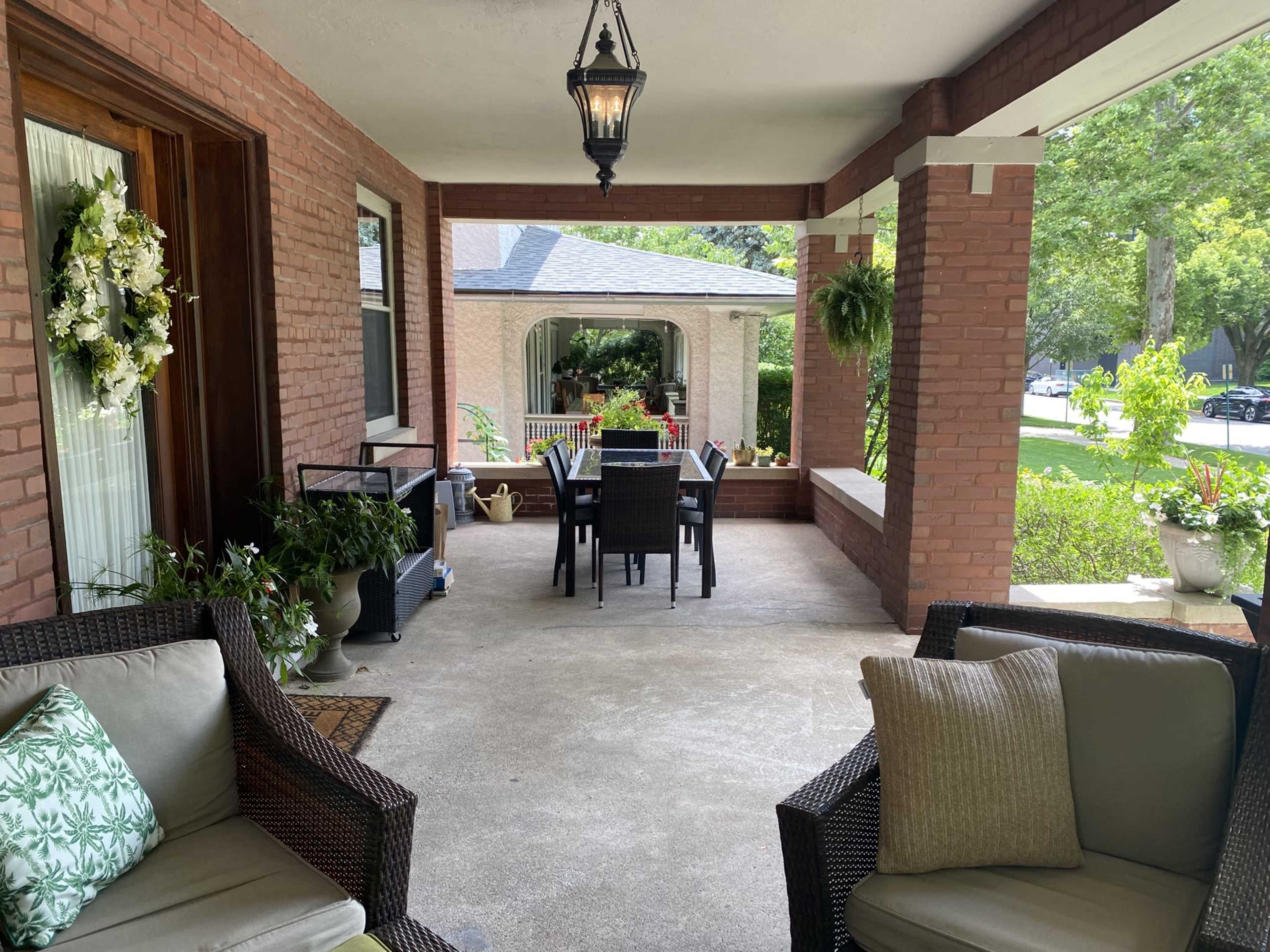 The image shows a covered porch featuring a seating area with wicker chairs and a dining table set up for four, surrounded by greenery and decorative plants.