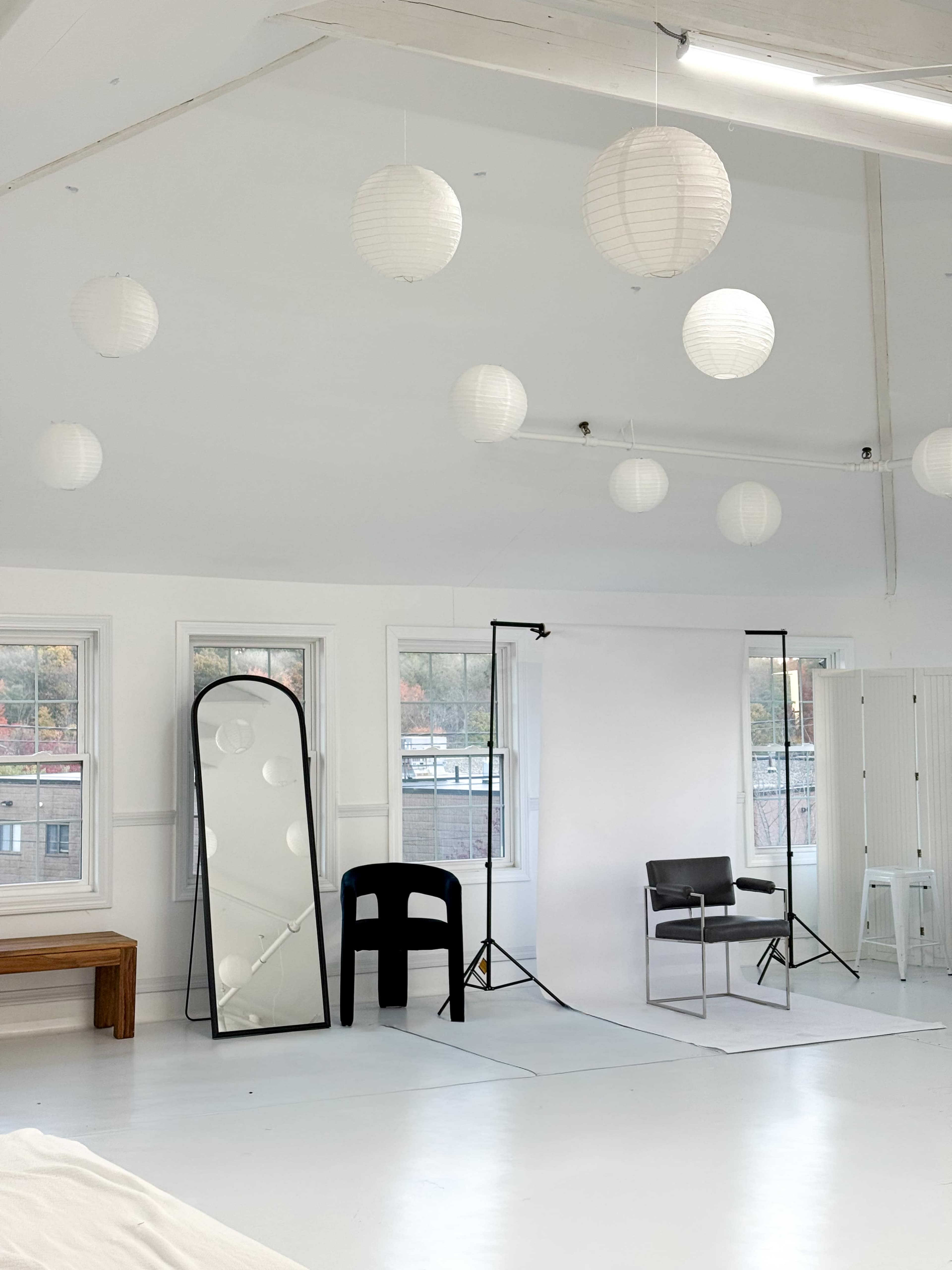 The image shows a bright, minimalist studio space with white walls, a large mirror, a black chair, and a backdrop arranged for photography, along with decorative paper lanterns hanging from the ceiling.