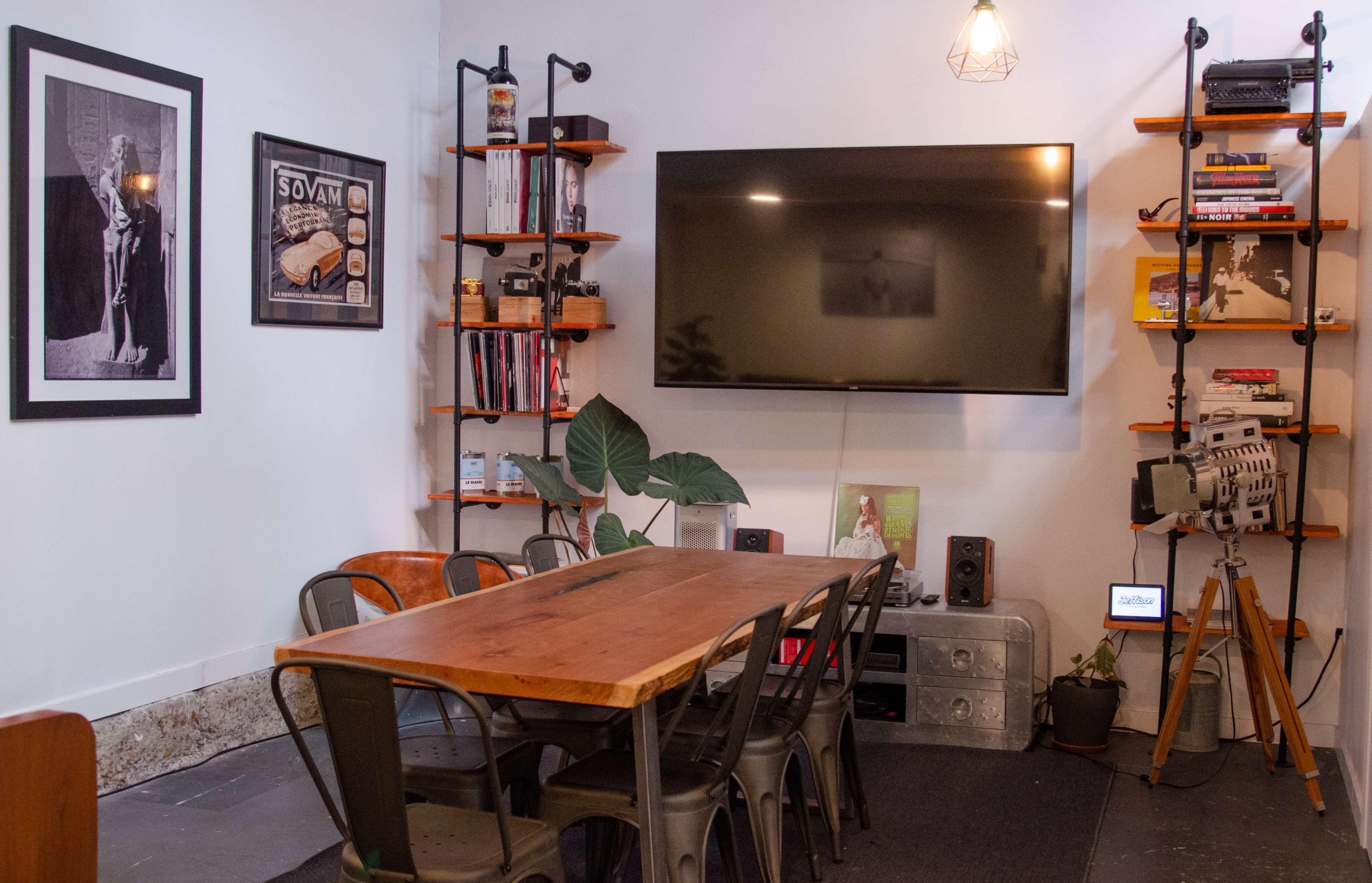 A modern dining area features a wooden table surrounded by metal chairs, with a large television mounted on the wall and bookshelves on either side, displaying various items and decorations.