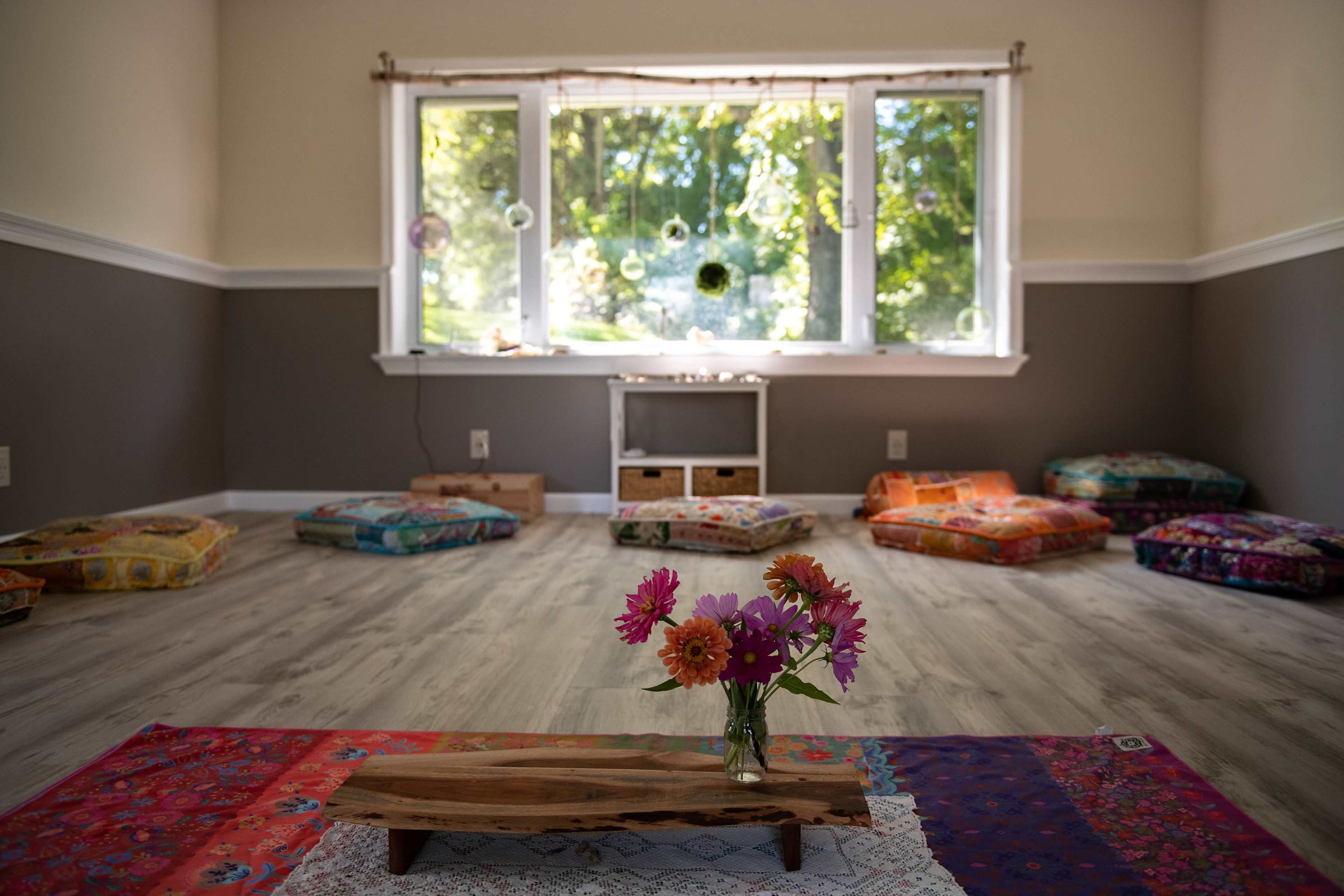 A bright, tranquil yoga studio features a wooden table with a floral vase at its center and colorful cushions arranged around the perimeter.