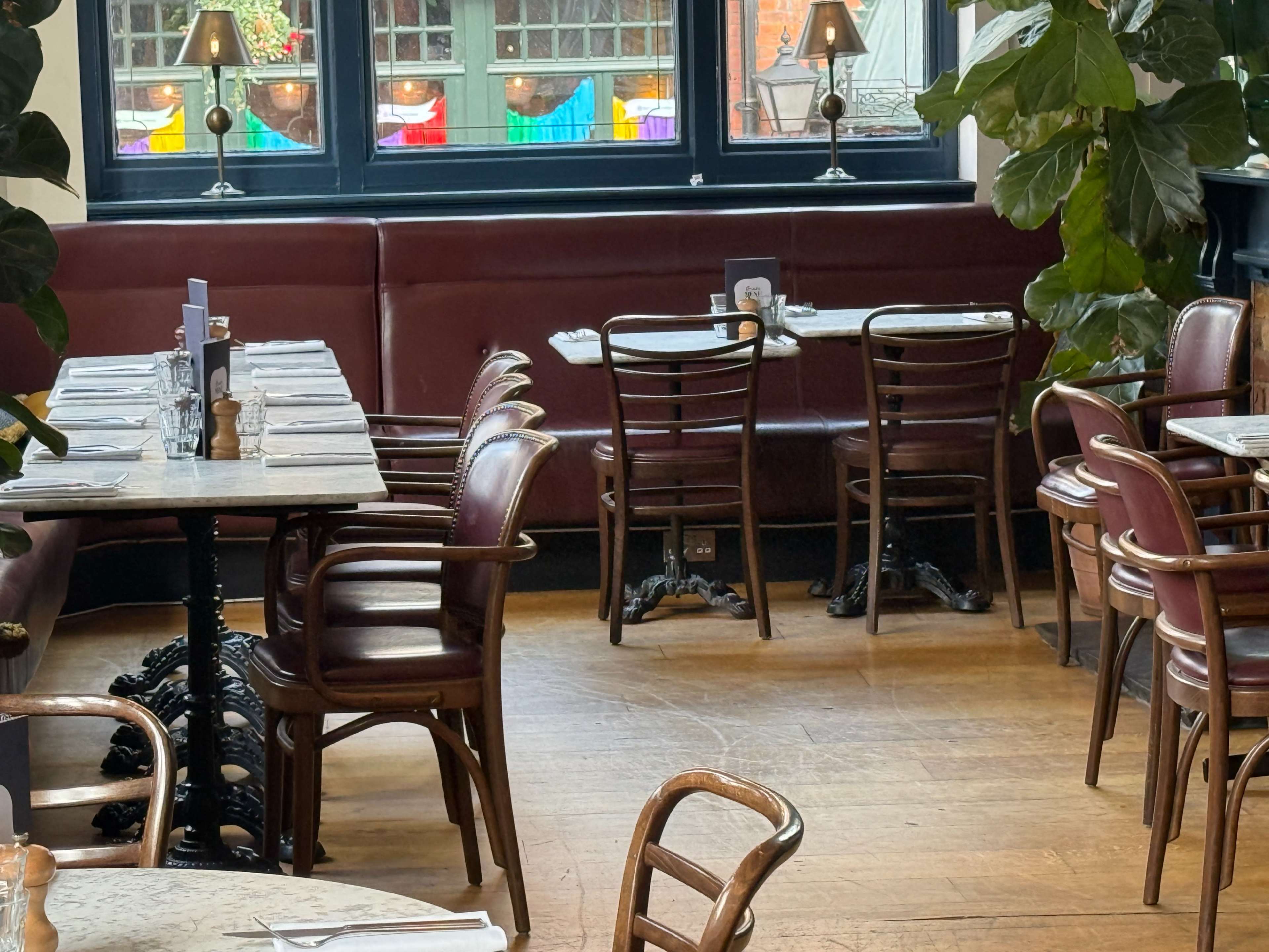 The image shows a restaurant interior with wooden tables and red upholstered chairs, complemented by potted plants and soft lighting from table lamps.