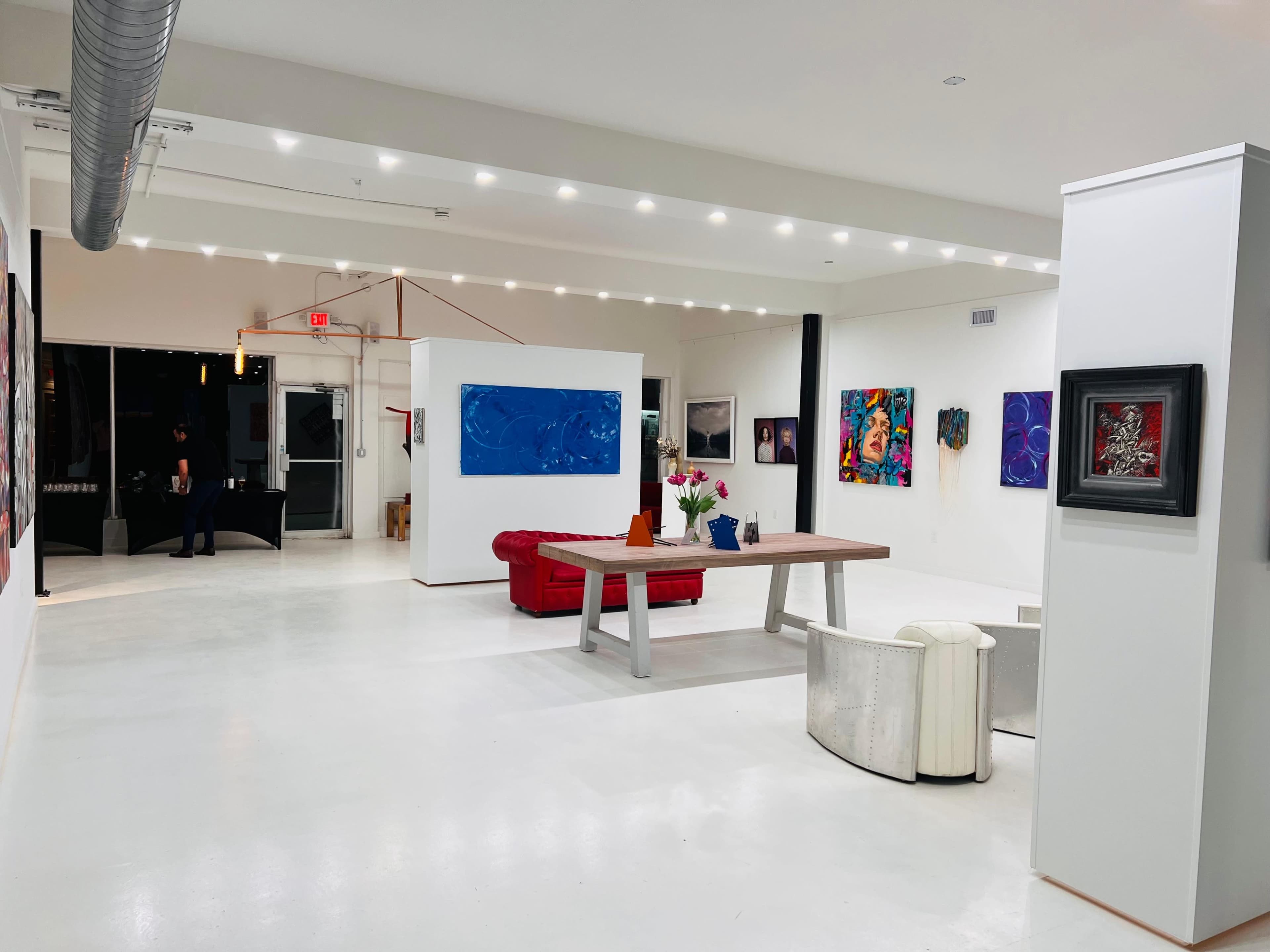 The image shows a spacious contemporary art gallery featuring a red sofa, a wooden table, and various colorful artworks displayed on white walls.