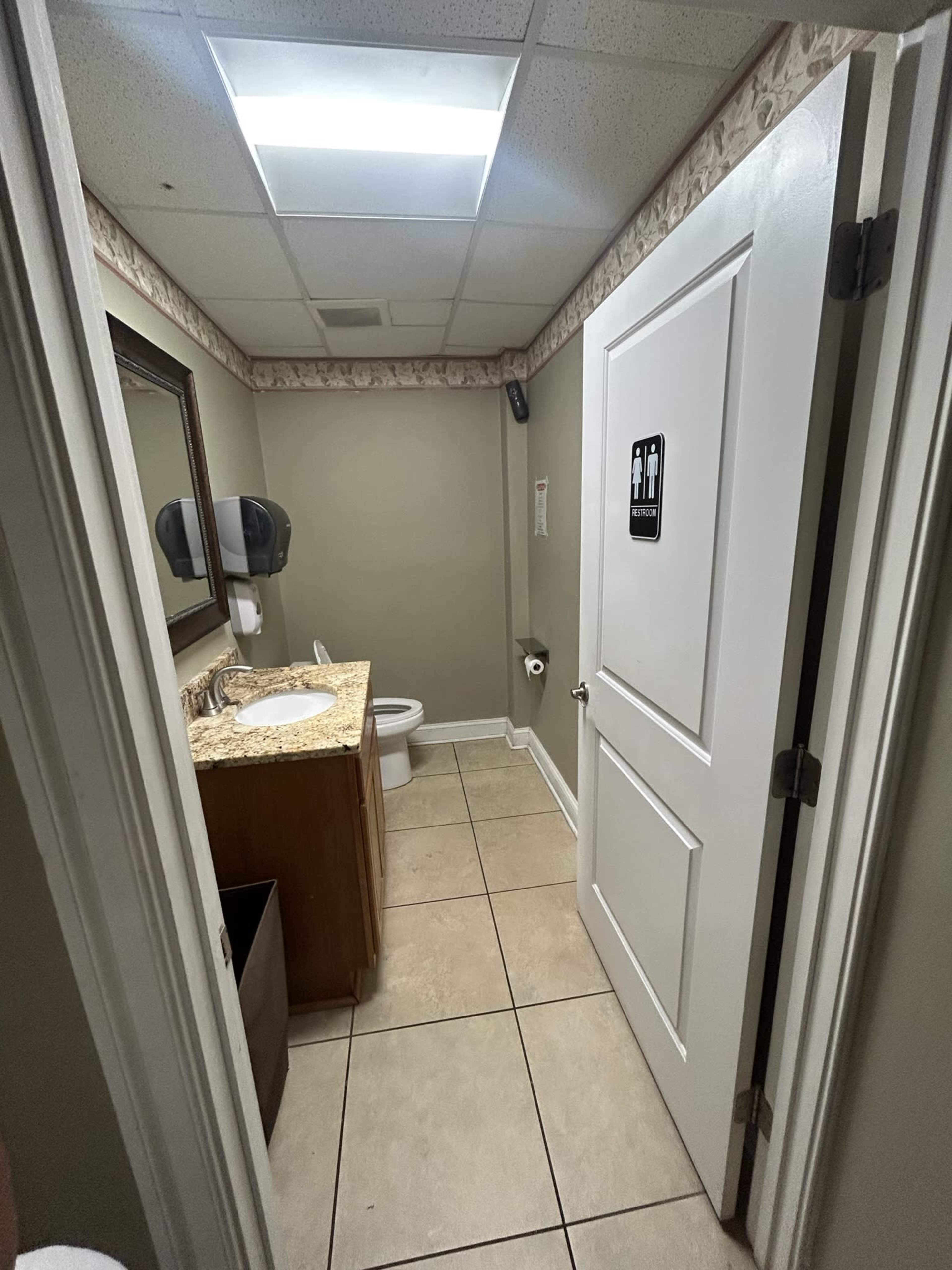 A small bathroom with a sink, mirror, toilet, and a door leading out, all within a tiled space.