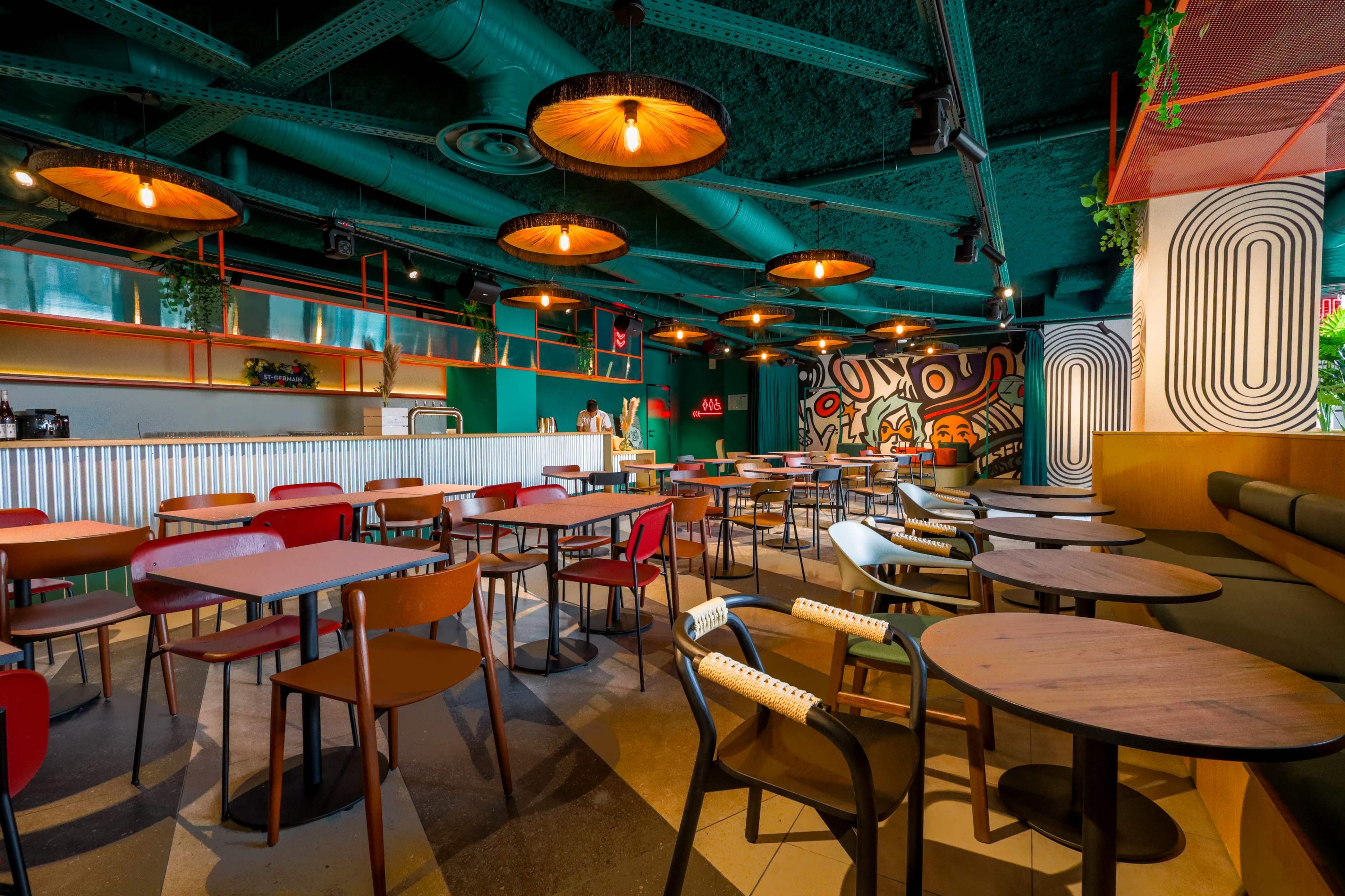The image shows a modern café interior with a variety of tables and chairs, colorful lighting fixtures, and a vibrant mural on the wall.