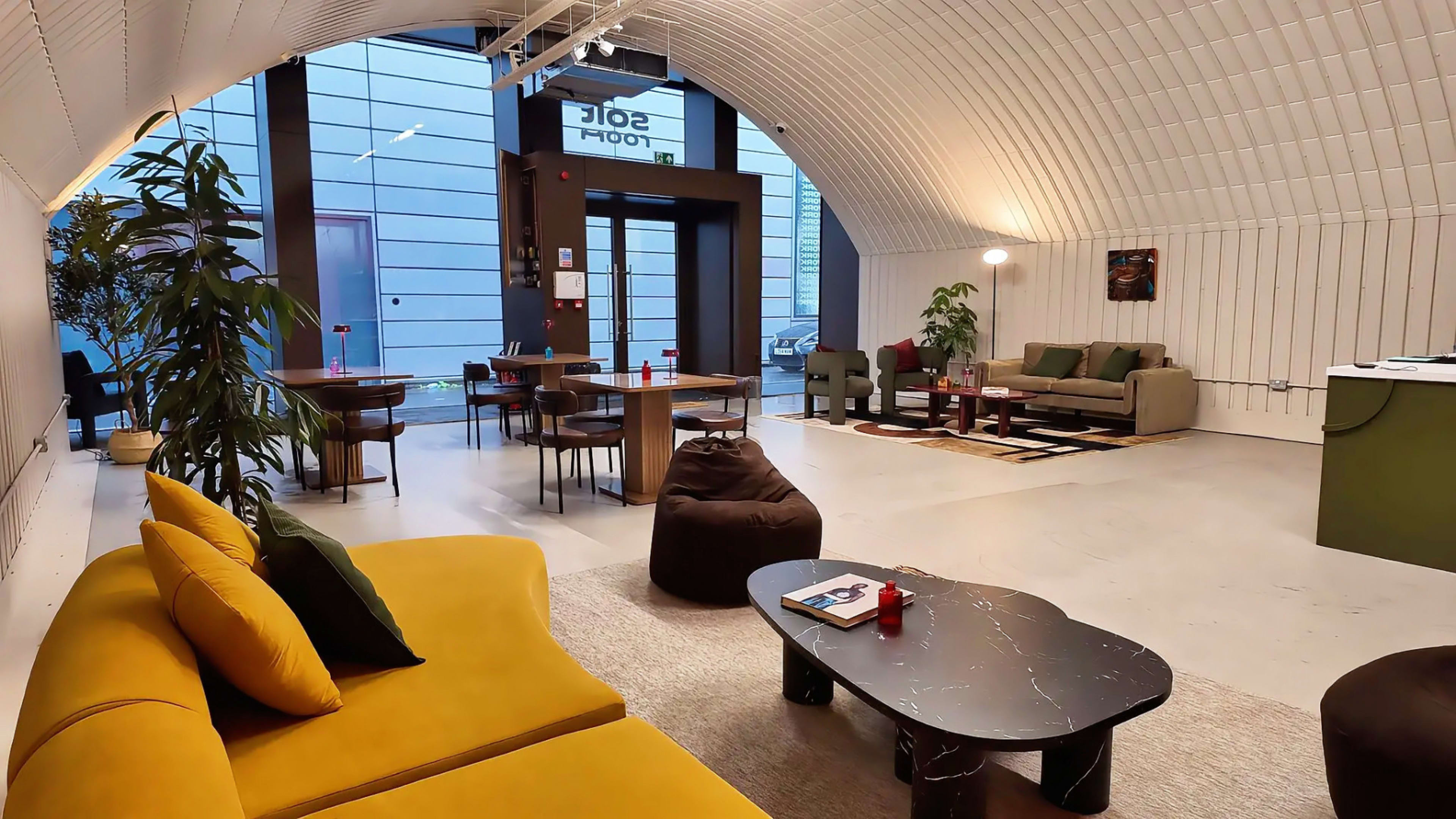 The image shows a modern, open lounge area with a curved ceiling, featuring yellow sofas, green seating, wooden tables, and a welcoming entrance.