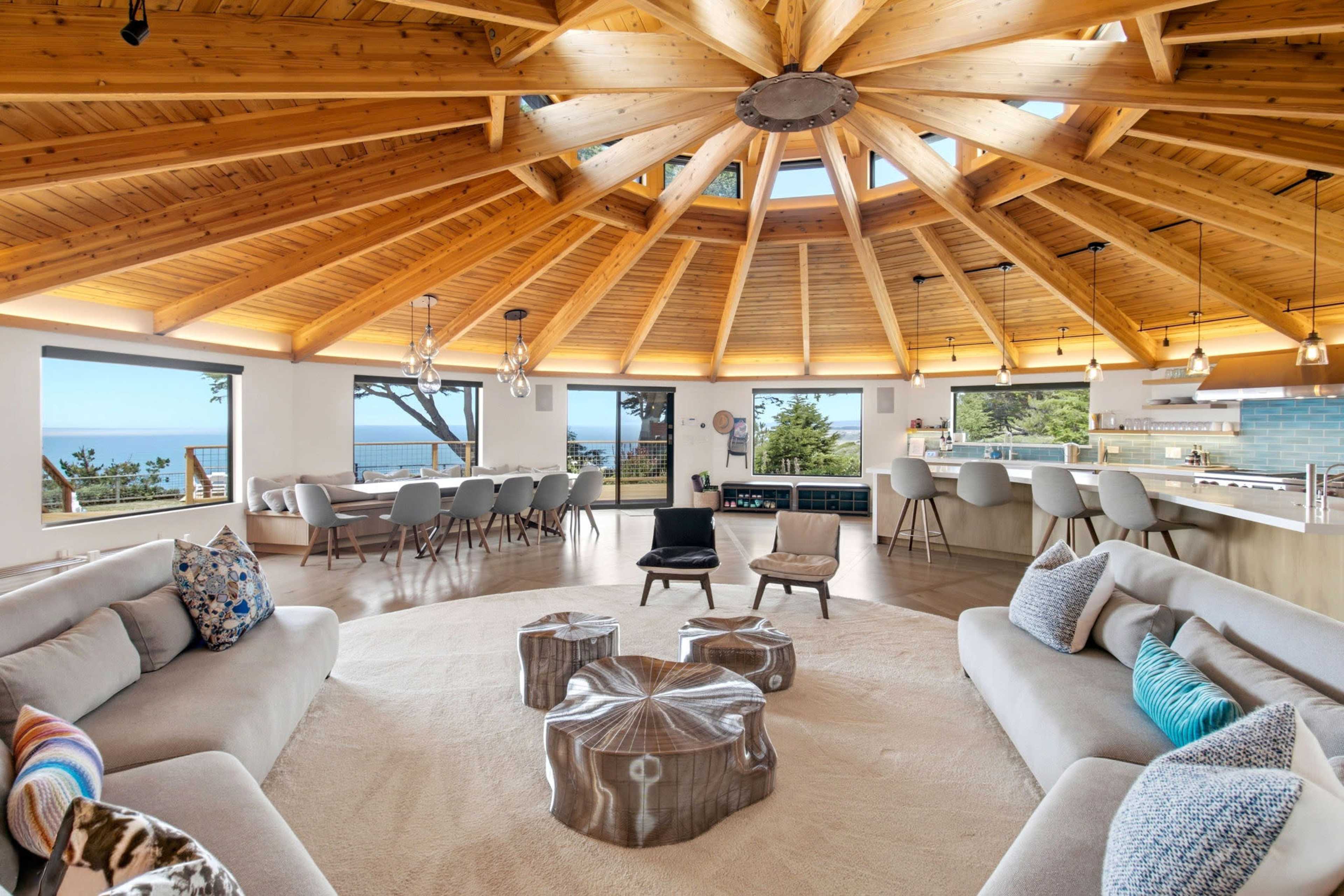 The interior of a circular-shaped living space features a wooden ceiling with skylights, surrounded by modern furniture and a view of the ocean.