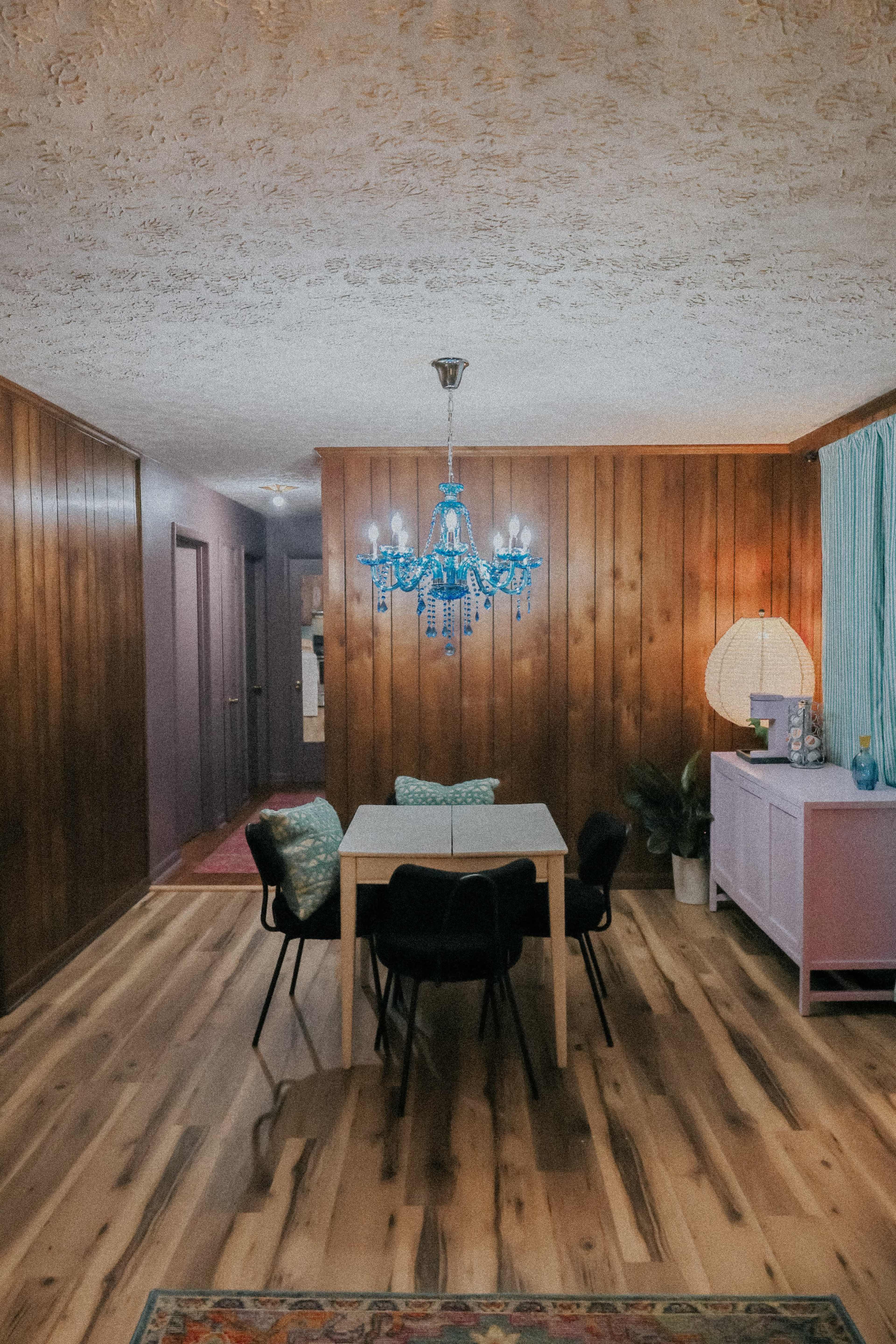 The image shows a dining area with a table and four chairs under a blue chandelier, surrounded by wooden walls and decorated with a floor rug.