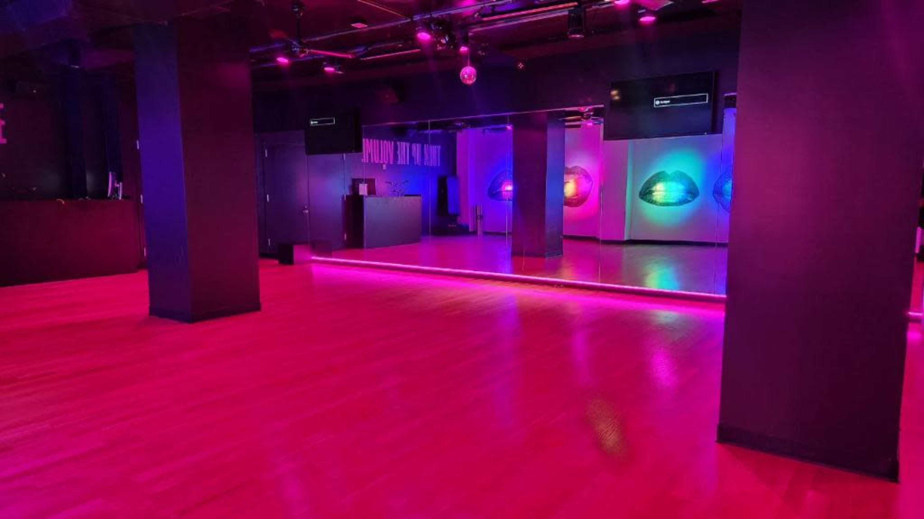 The image shows a dimly lit dance studio featuring wooden floors, colorful lighting, and a mirrored wall.