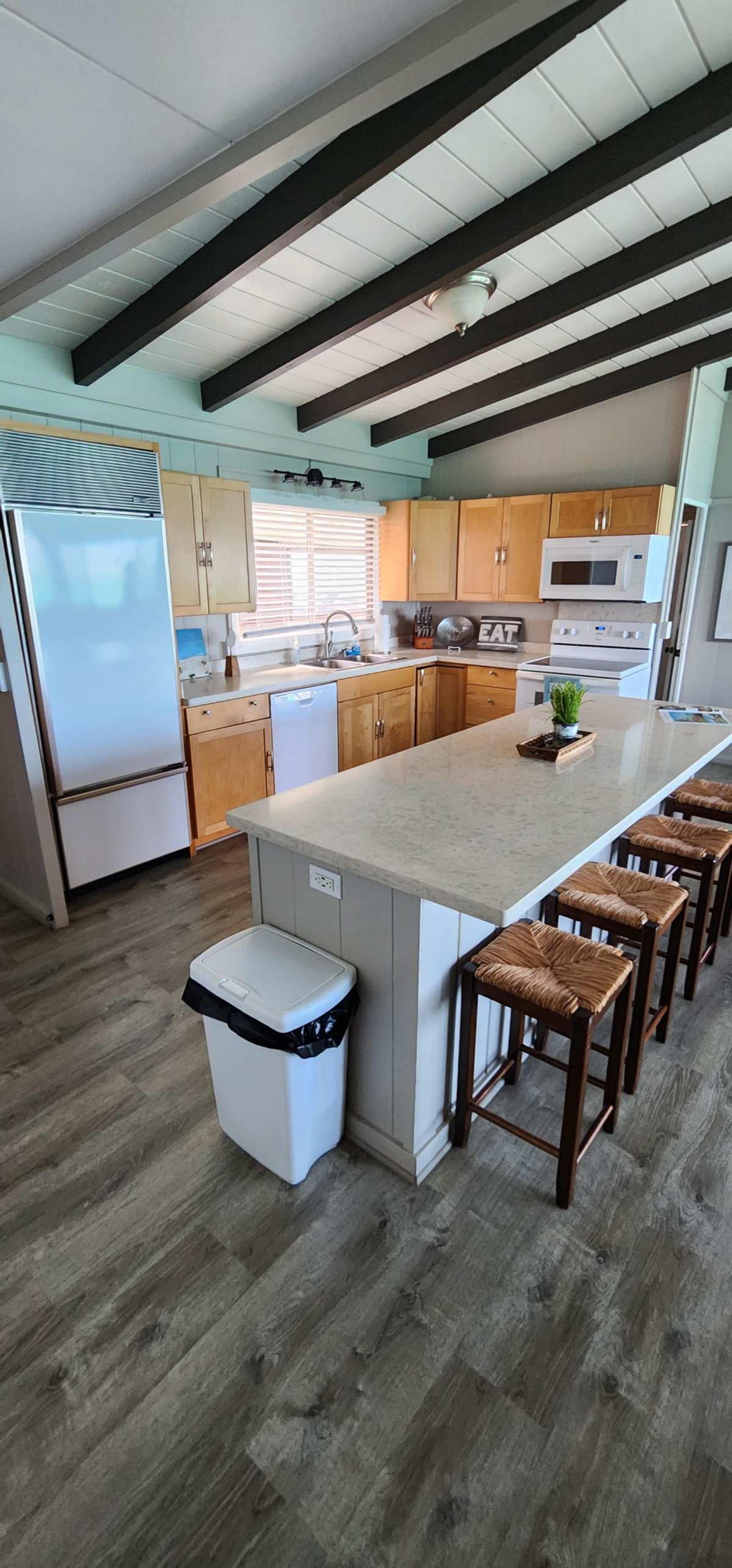 The image shows a modern kitchen with wooden cabinets, a large island with bar stools, and white appliances.