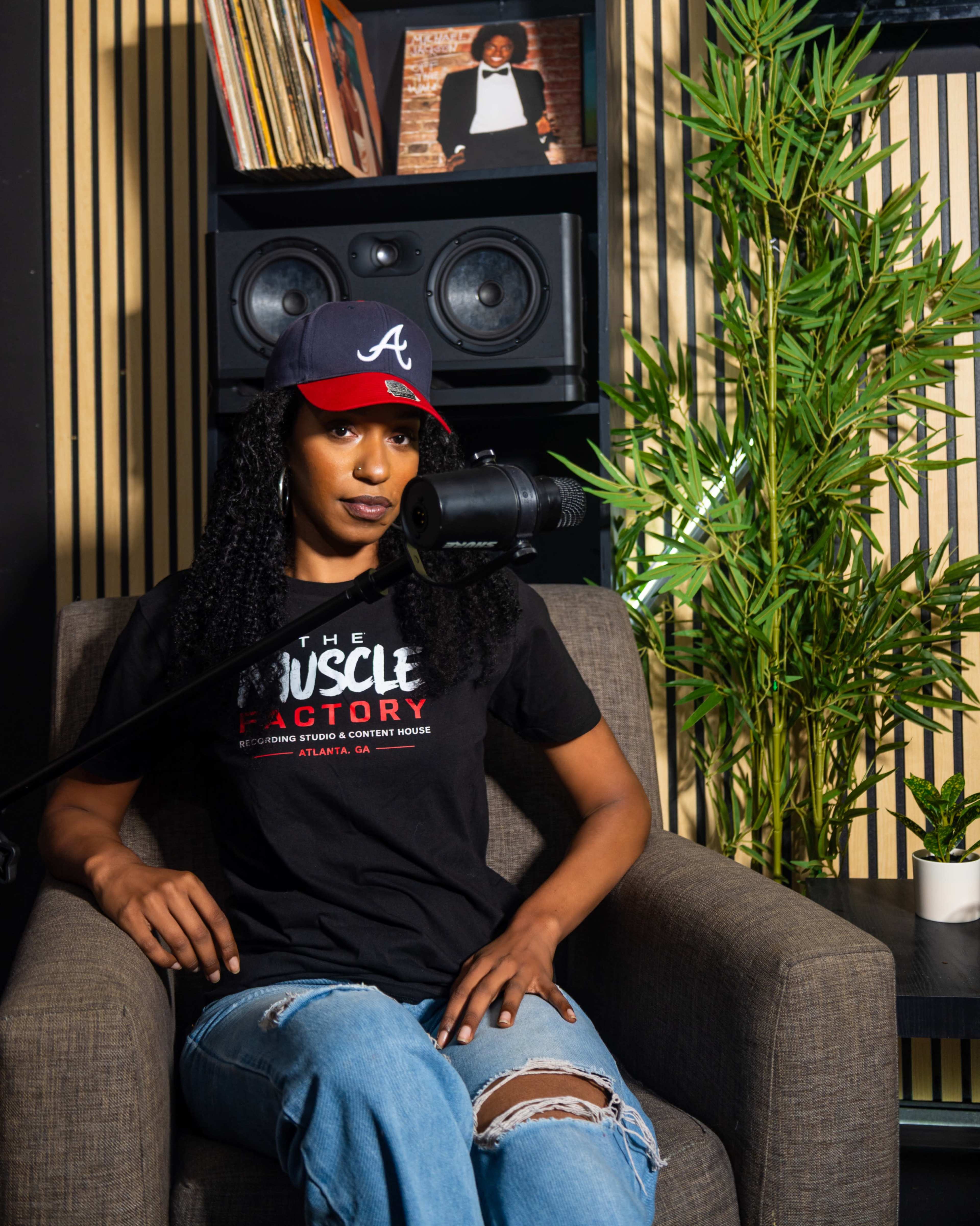 A person wearing an "Atlanta" baseball cap sits in a chair with a microphone in front of them, surrounded by plants and speakers in a stylish room.