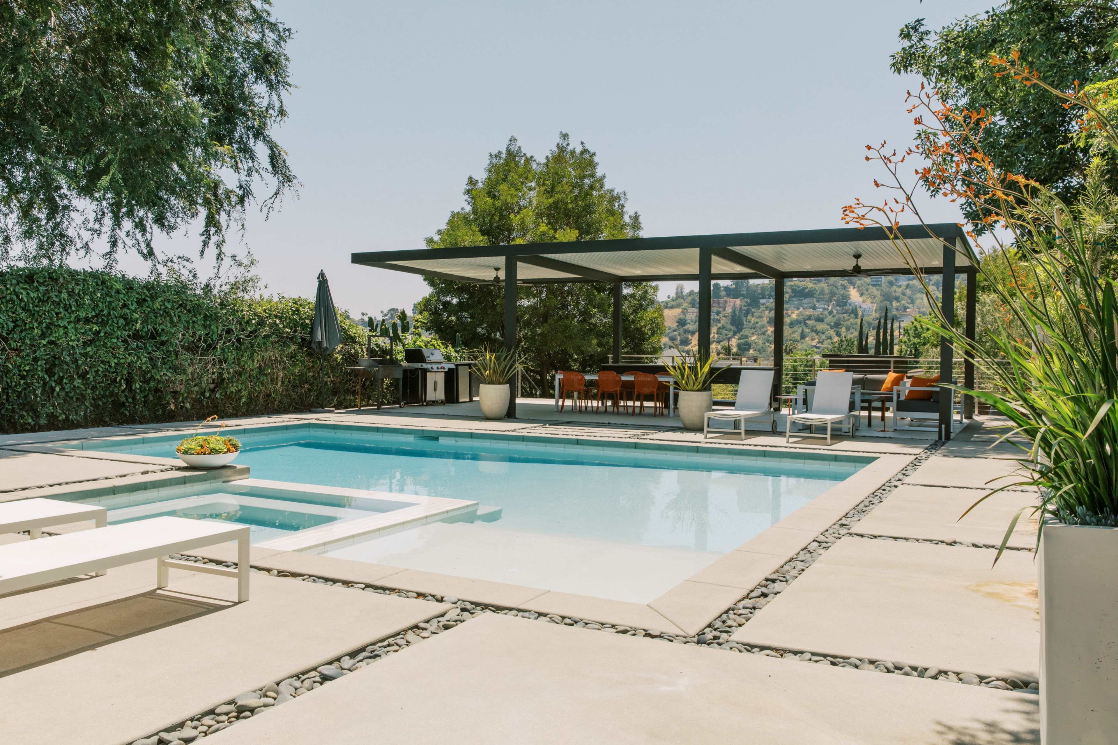 A modern backyard features a swimming pool surrounded by concrete decking, with a covered seating area and outdoor dining set in the background.