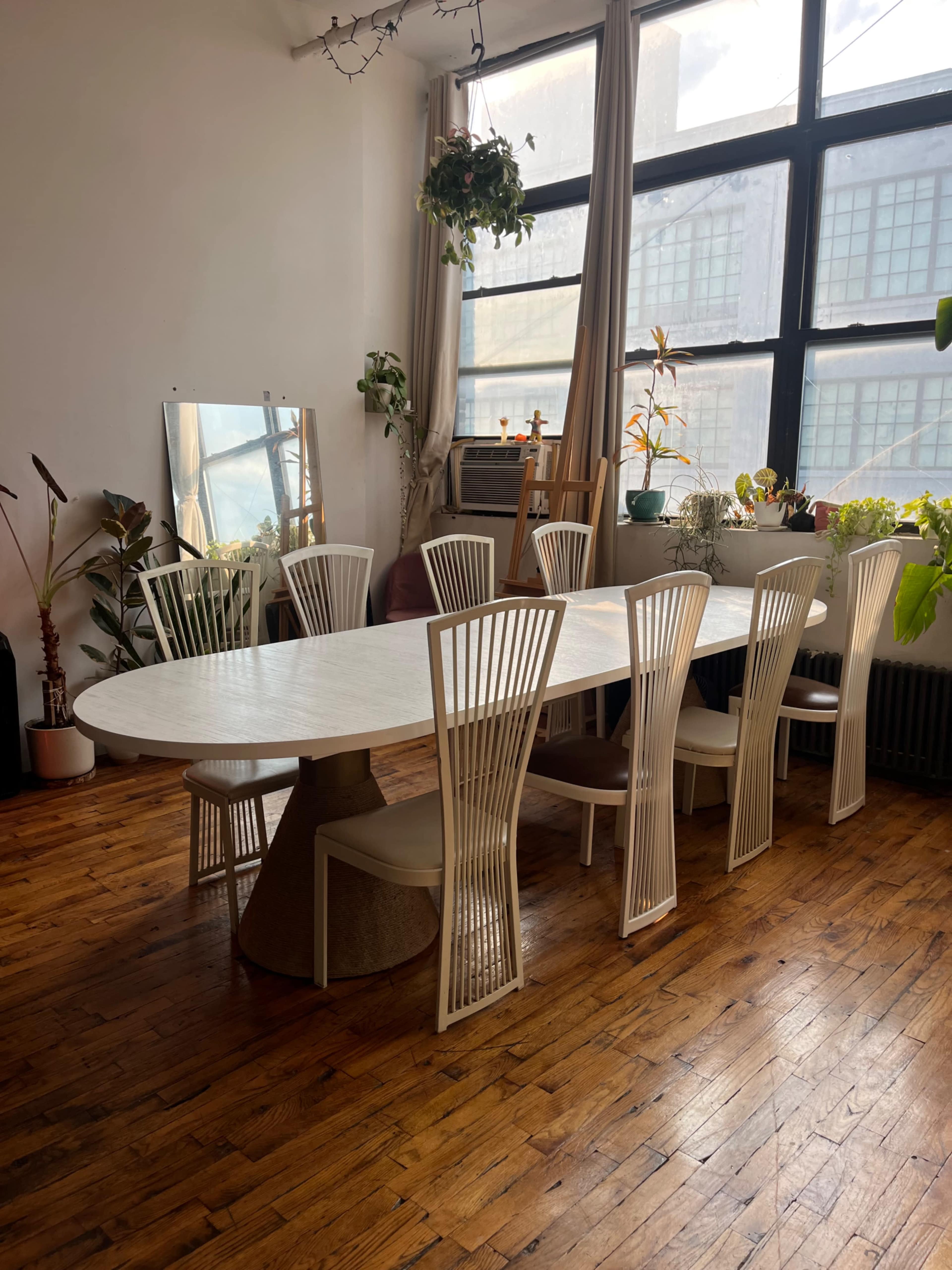 The image shows a long, oval dining table surrounded by white chairs in a sunlit room with plants and large windows.