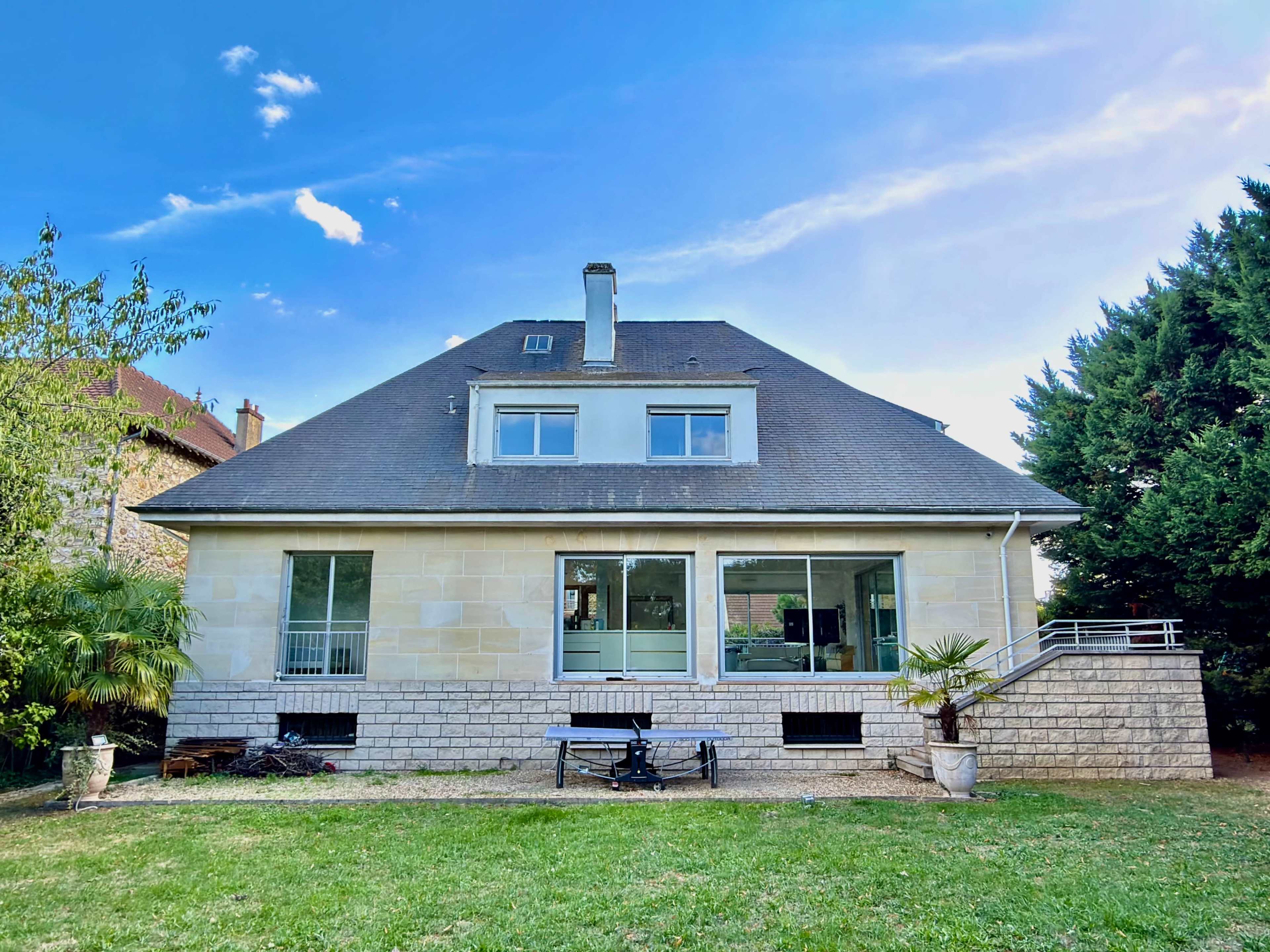 The image shows a two-story house with a sloped roof, large windows at the back, and a grassy yard surrounded by trees.