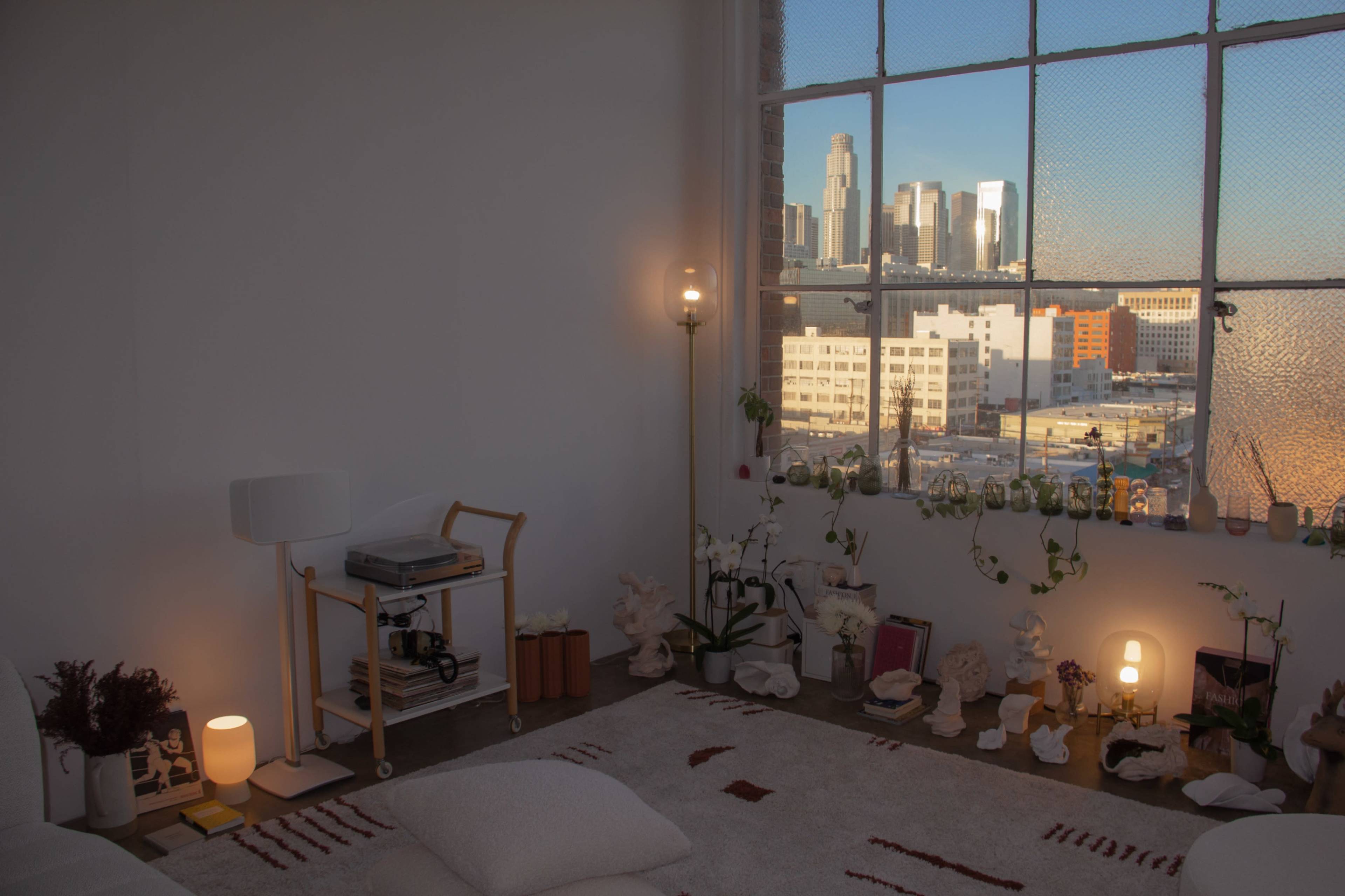 The interior features a cozy seating area with a lamp, a cart with a record player, and large windows overlooking a cityscape.