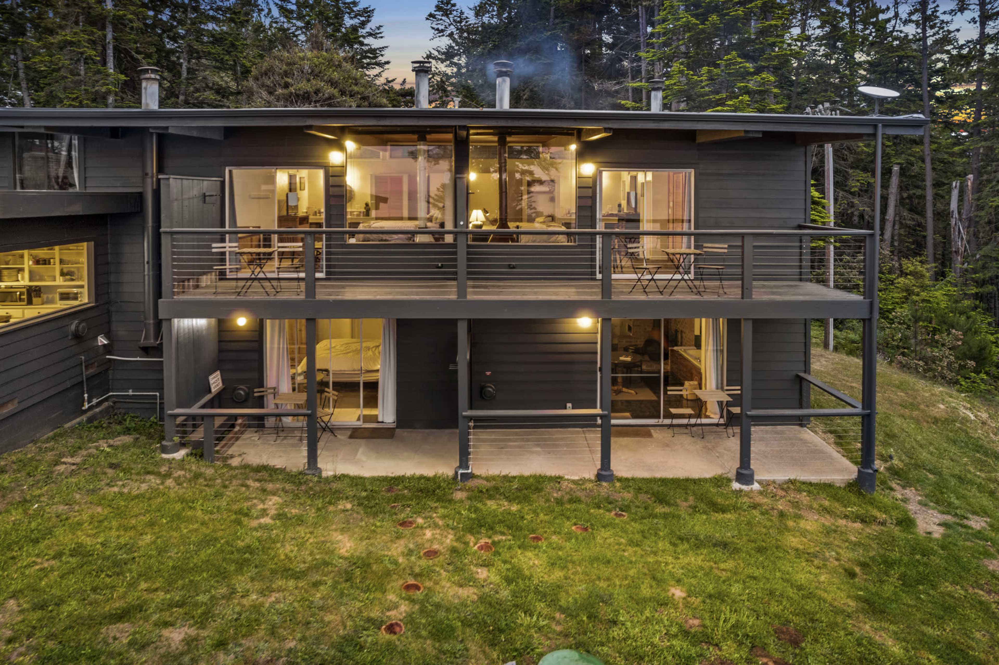 A modern two-story black house features large windows and a balcony overlooking a green lawn and trees.