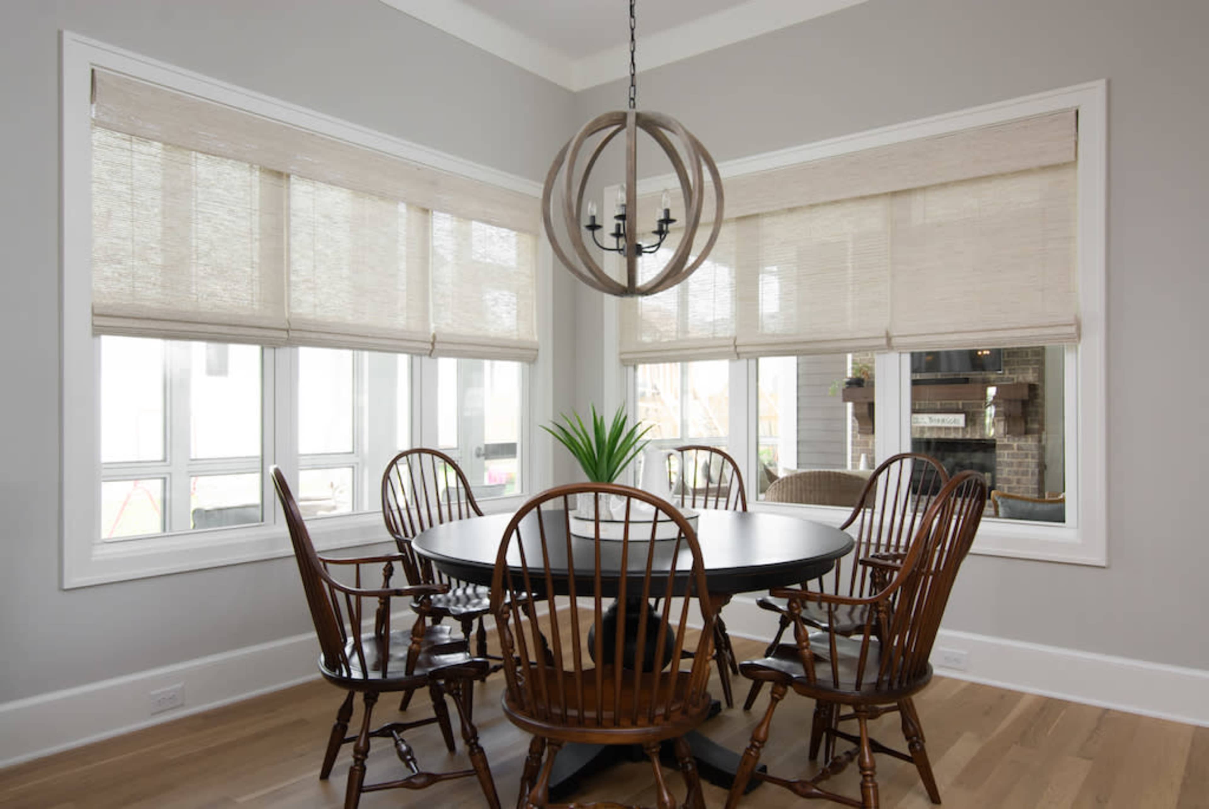 A circular wooden dining table surrounded by six wooden chairs is positioned in a bright room with large windows and light-colored walls.