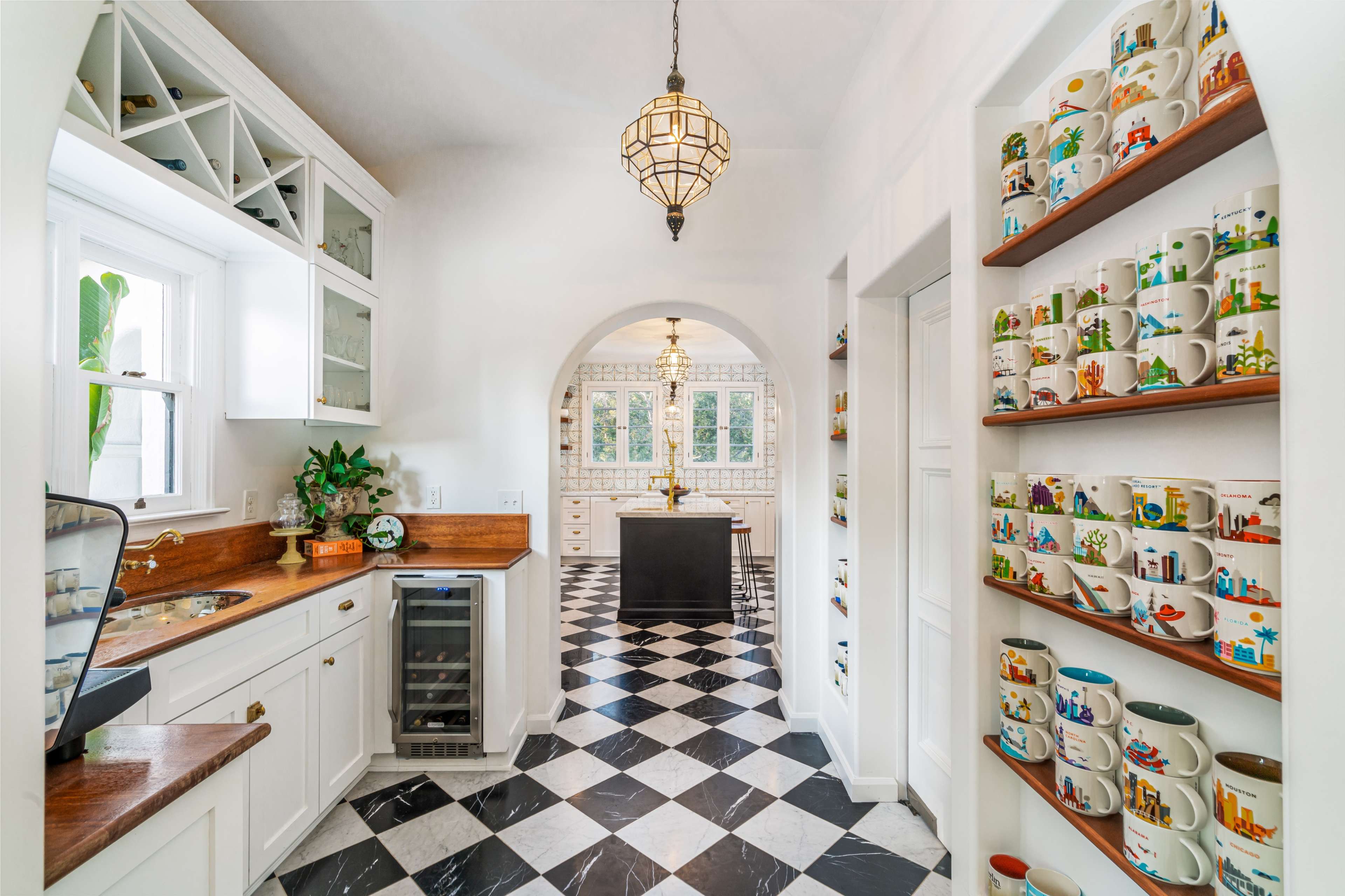 A bright kitchen features white cabinetry, a black and white checkered floor, and shelves lined with colorful mugs.