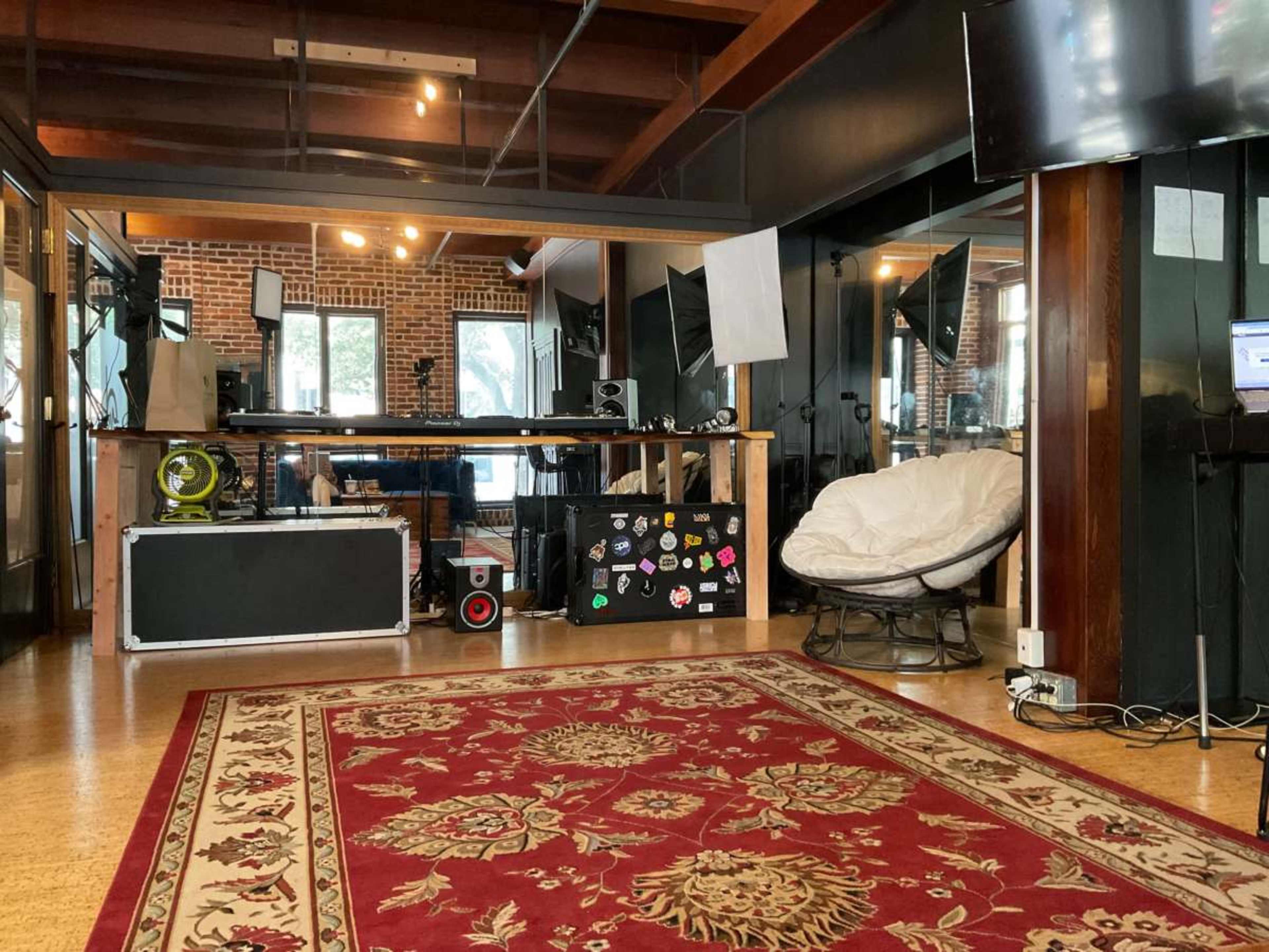 The image shows a bright, open space with wooden beams, a large area rug, and various workstations featuring monitors and black furniture.