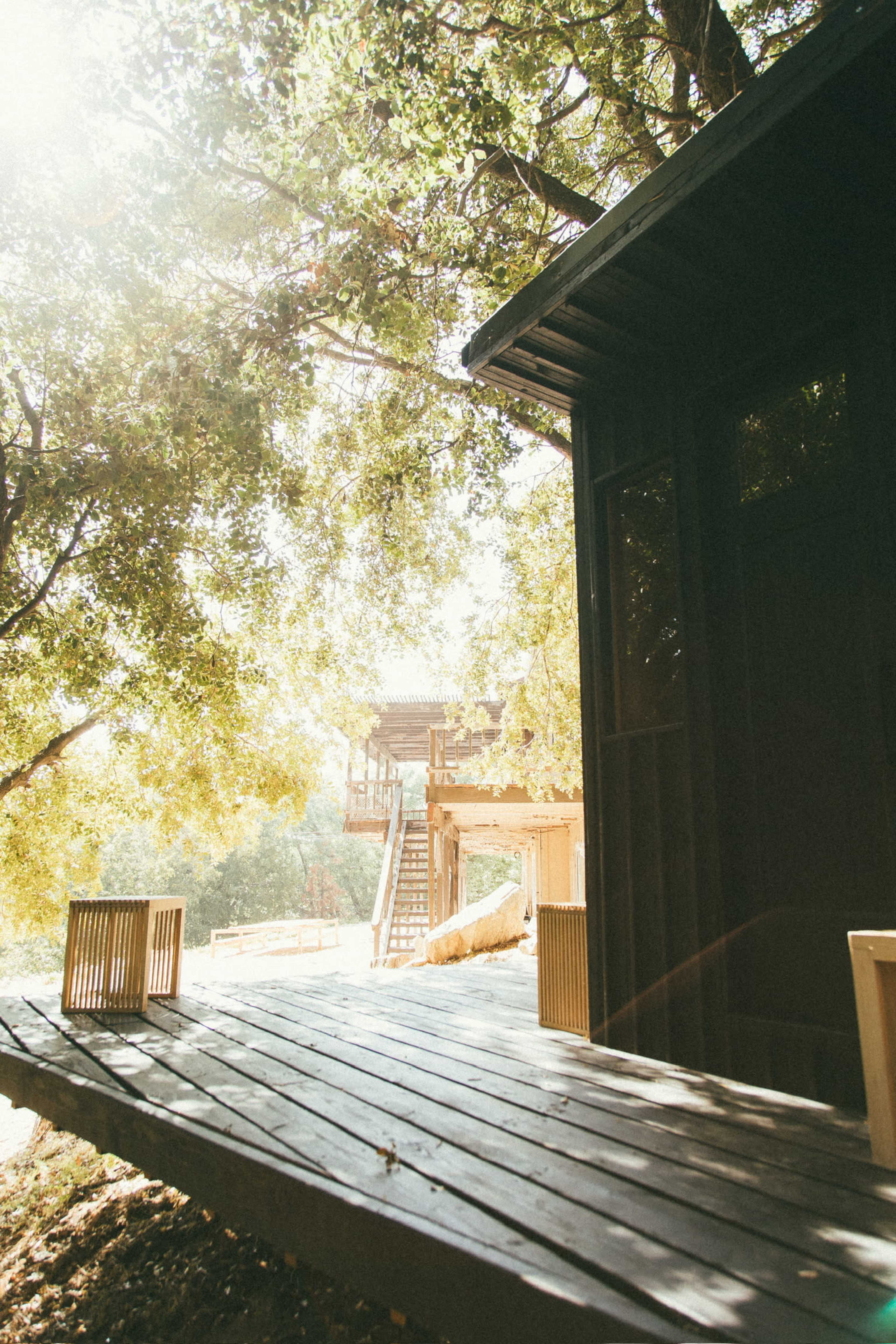 A wooden deck leading to a small cabin surrounded by trees, with a distant view of a staircase and another wooden structure.