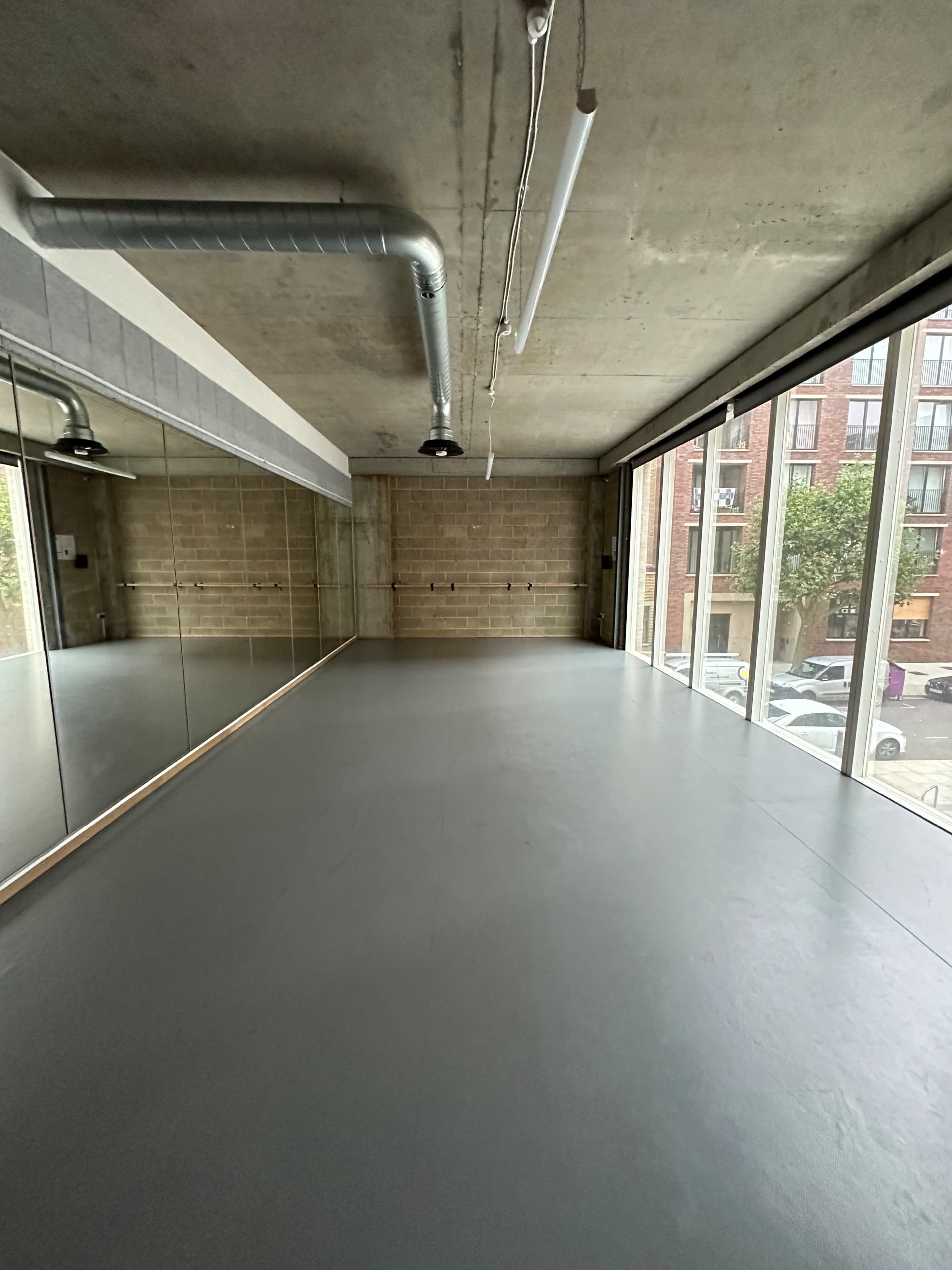 An empty room with a grey floor, large windows, and a mirrored wall, featuring exposed concrete walls and industrial lighting.